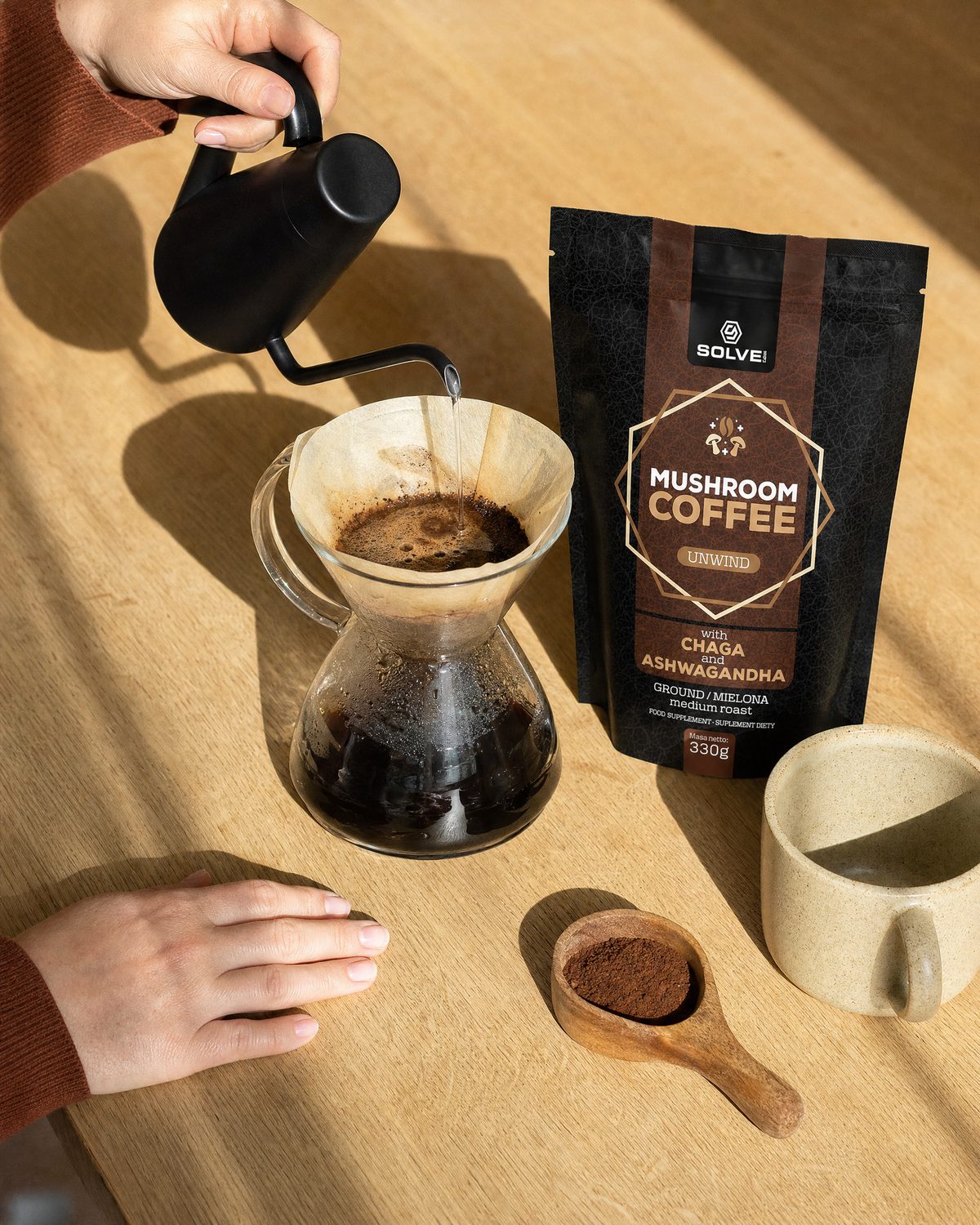Hands pouring water from a kettle over a pour-over carafe, a bag of Mushroom Coffee with Chaga and a stoneware cup beside it