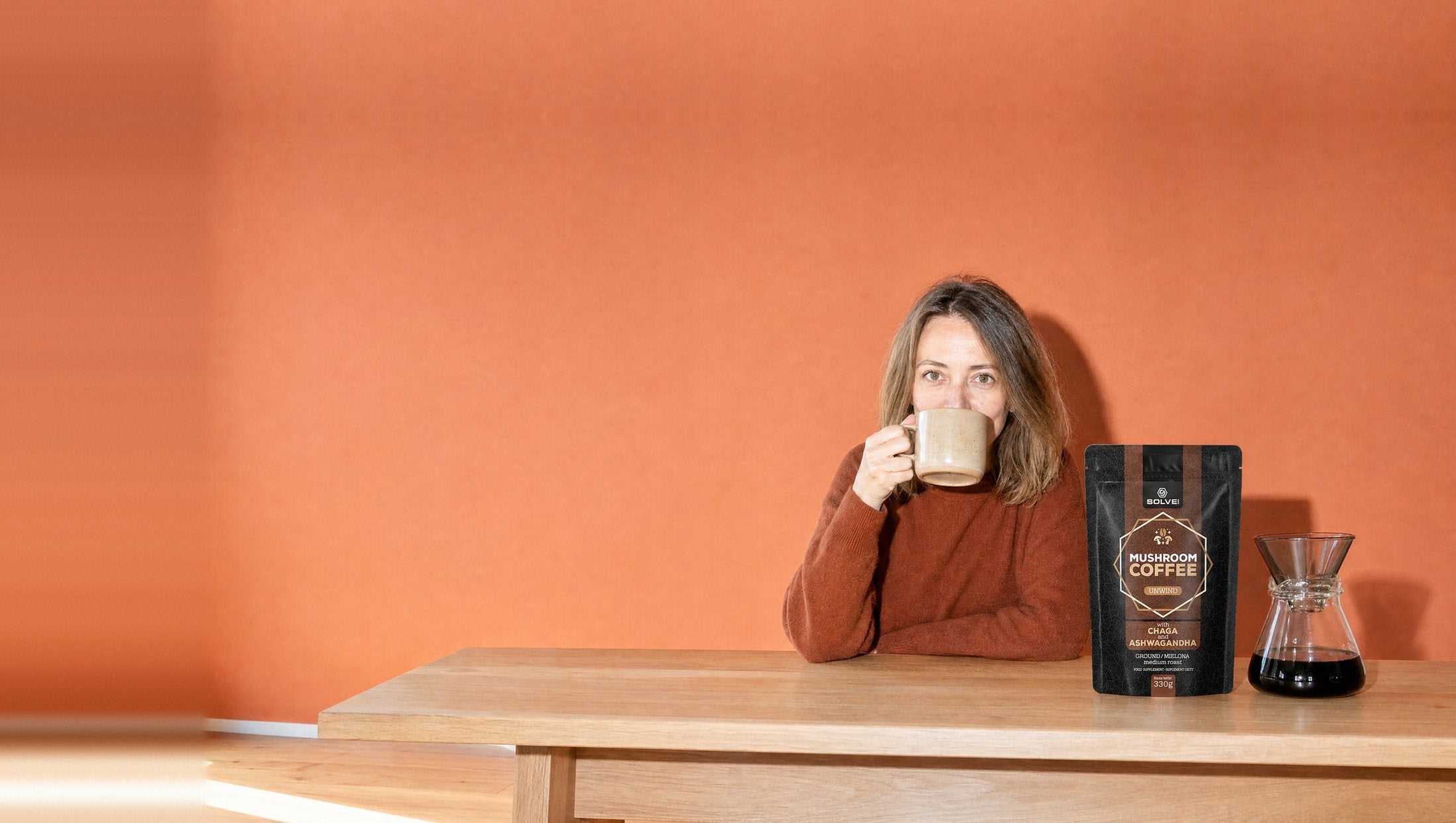A woman at a table in front of a terracotta wall drinking coffee from a stoneware cup, a bag of Solve Labs Mushroom Coffee with Chaga and a pour-over carafe beside her