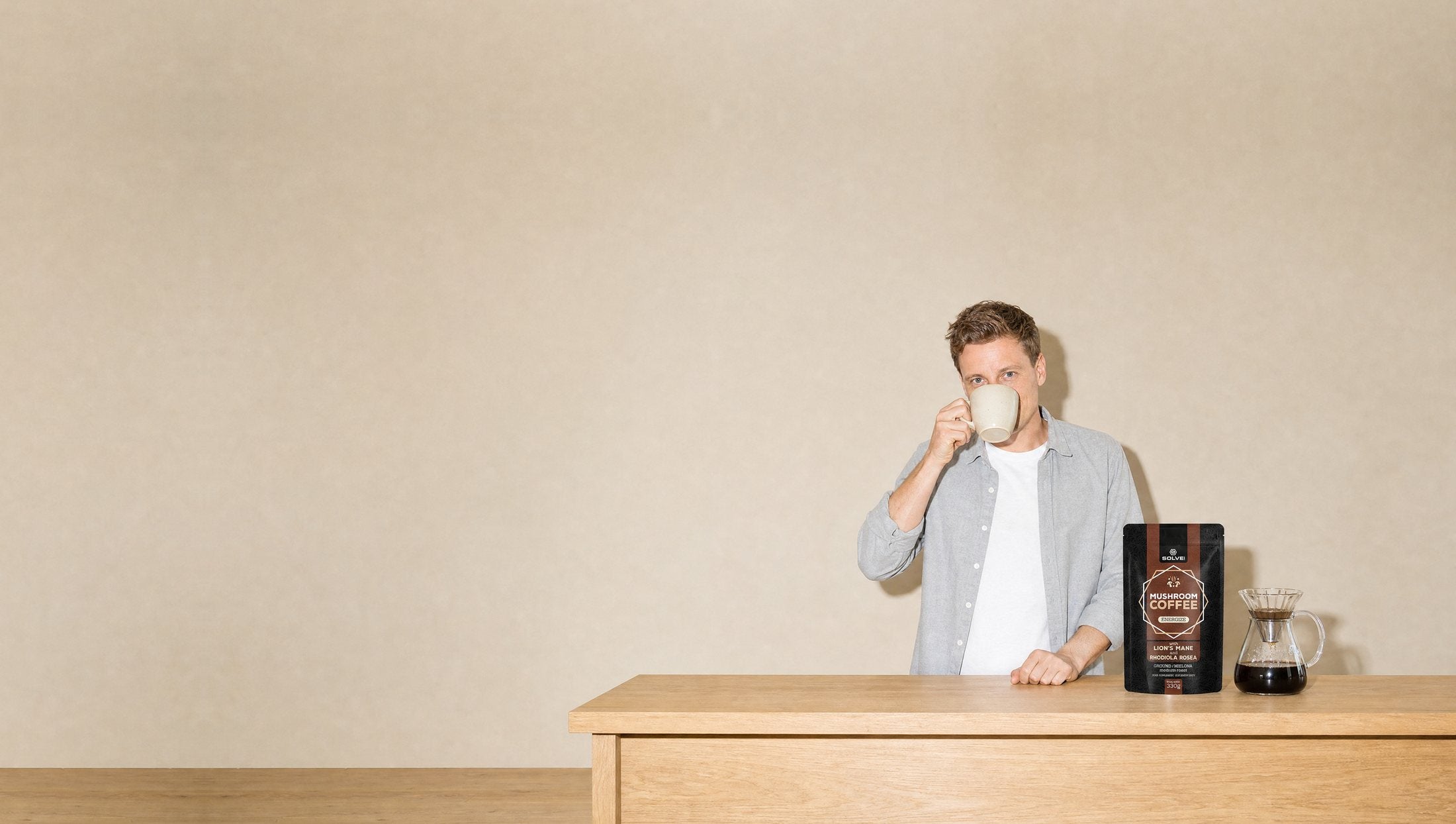 A man in a bright kitchen drinking coffee from a stoneware cup, a bag of Solve Labs Mushroom Coffee with Lion’s Mane and a pour-over carafe on the worktop beside him