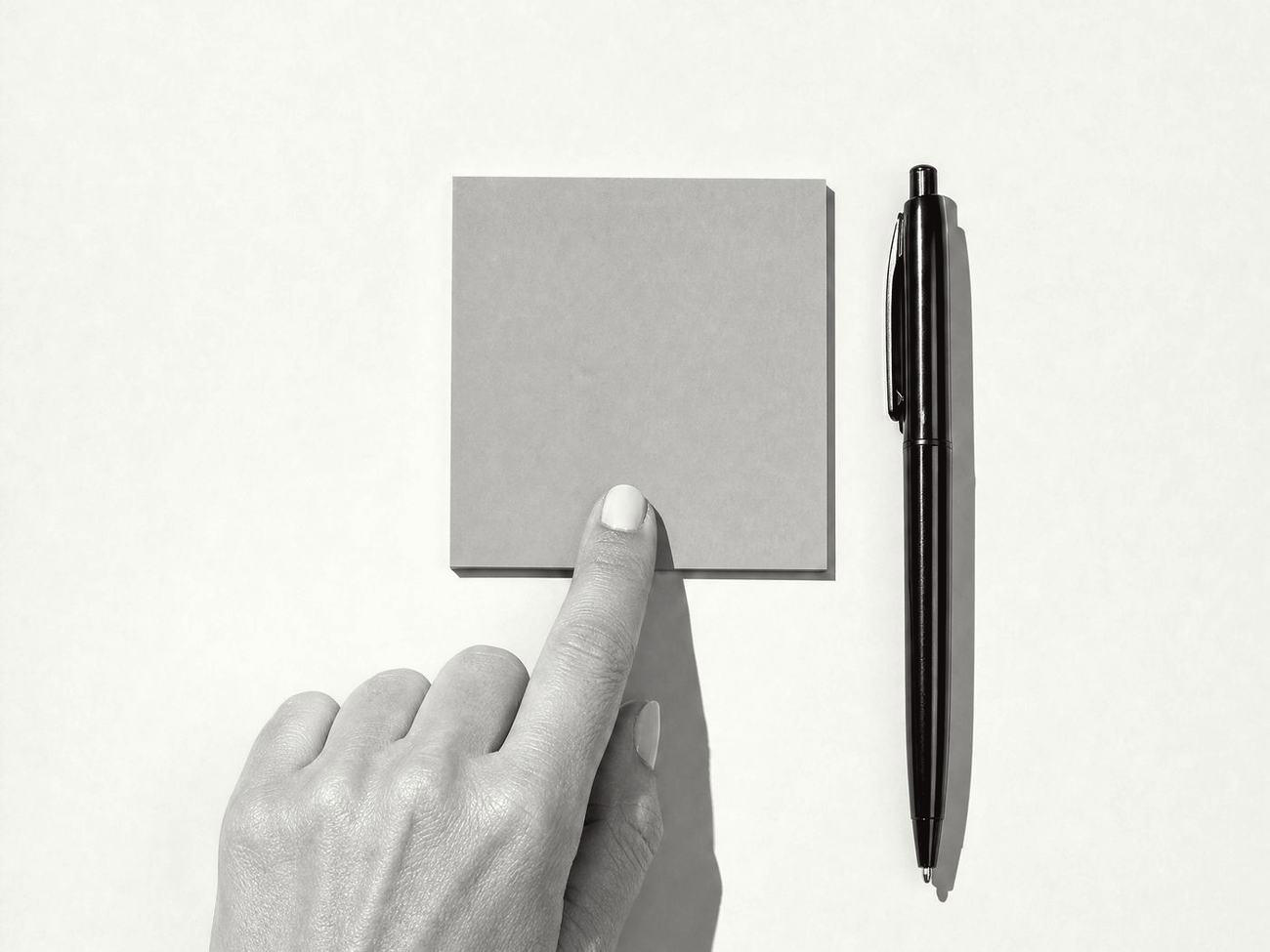 A fingertip pressing a single blank sticky note onto a desk, a pen beside it, in black and white