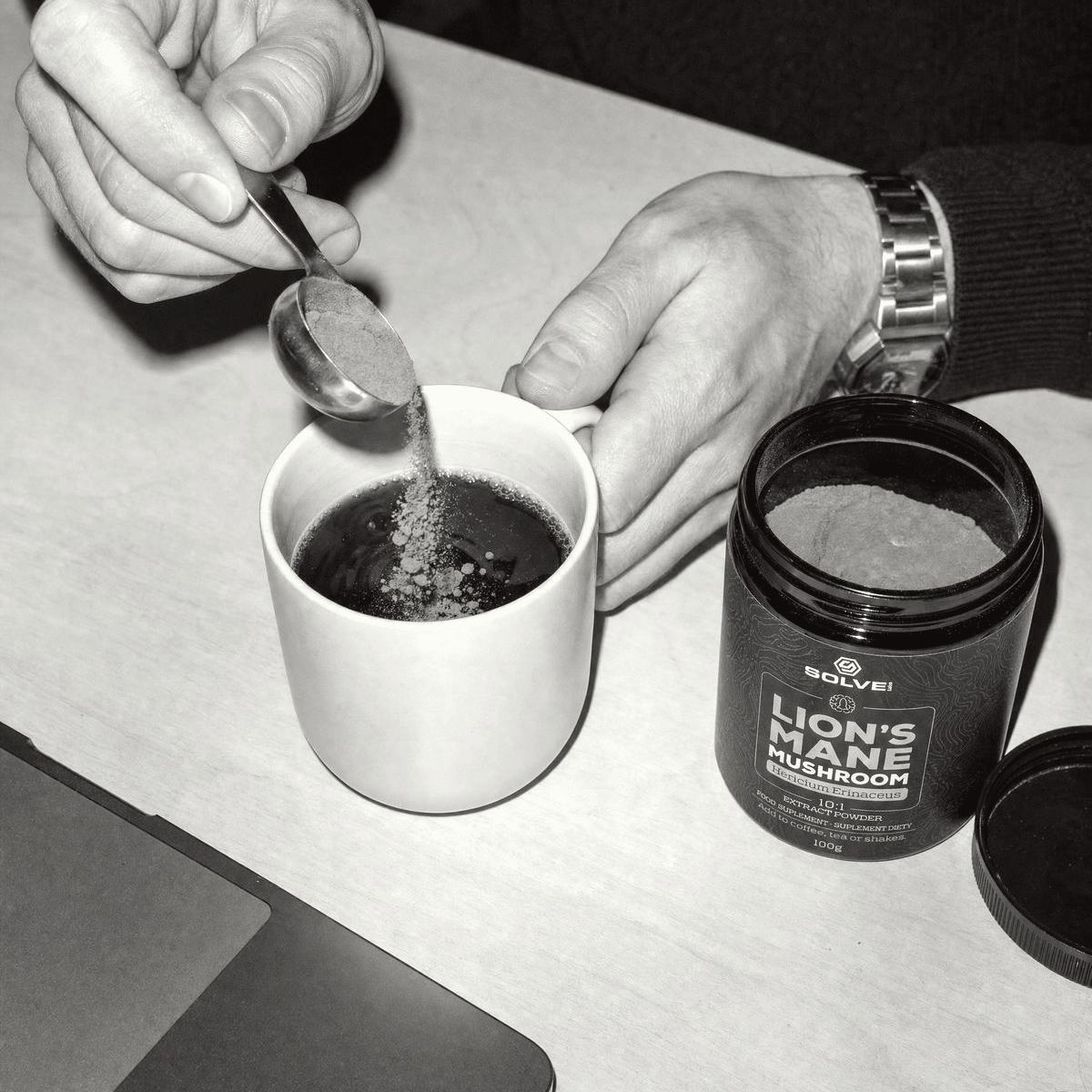 A hand tipping a spoon of Lion's Mane powder into a mug of black coffee on a desk, in black and white