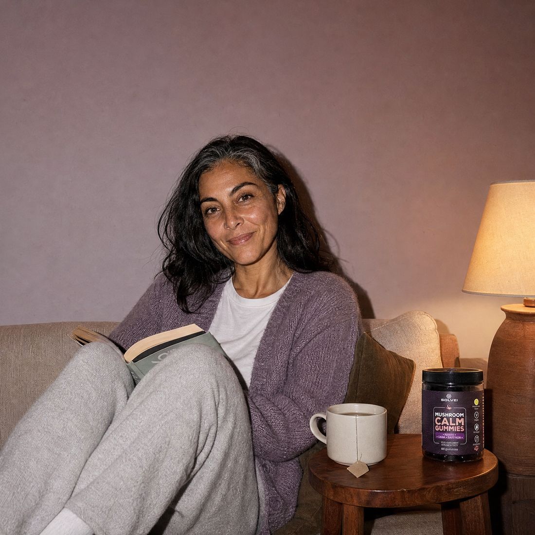 A woman curled up with a book on a low sofa beside a lit lamp, a mug and the jar of Calm Gummies on the side table