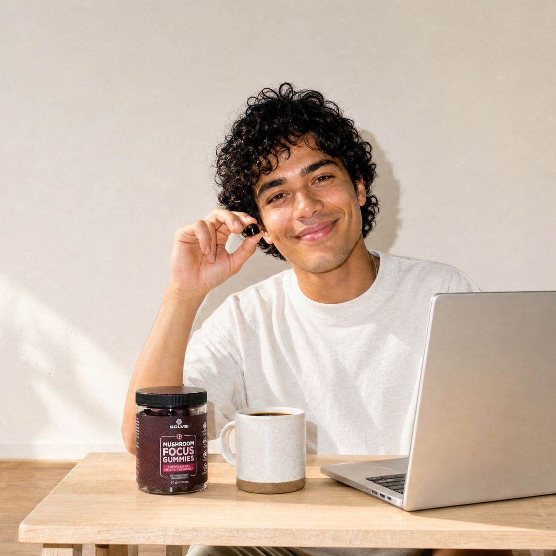 A young man at a sunlit desk with a laptop and a mug, holding up the jar of Focus Gummies