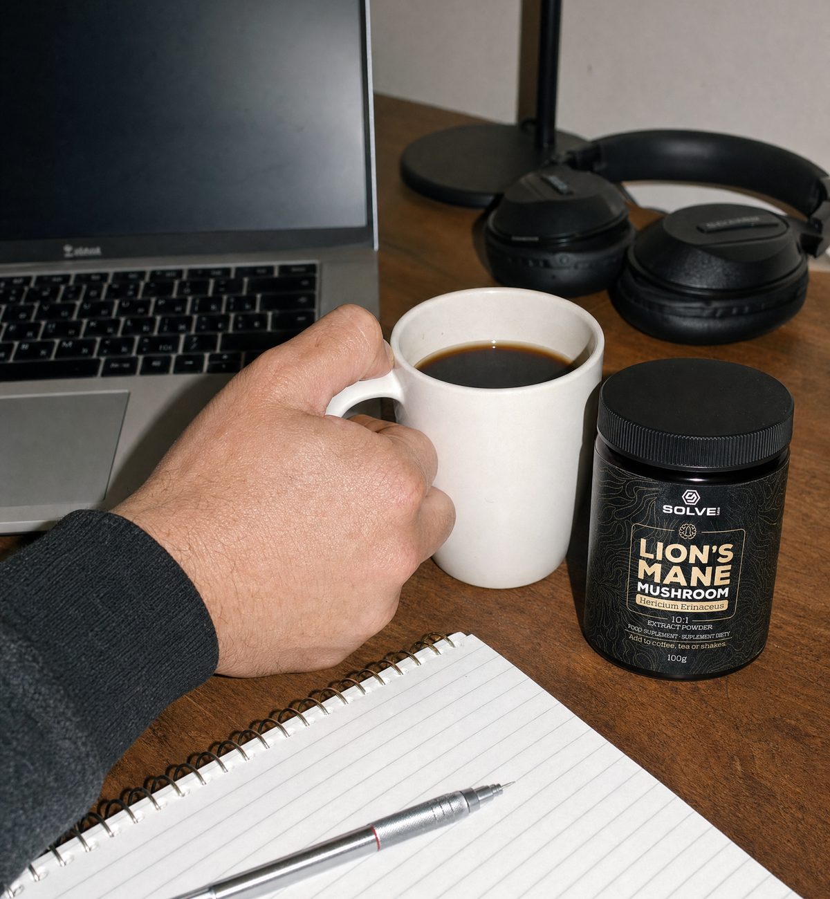 A desk seen from above: a laptop, headphones, a notebook and pen, a hand on a mug of black coffee, and a jar of Lion’s Mane