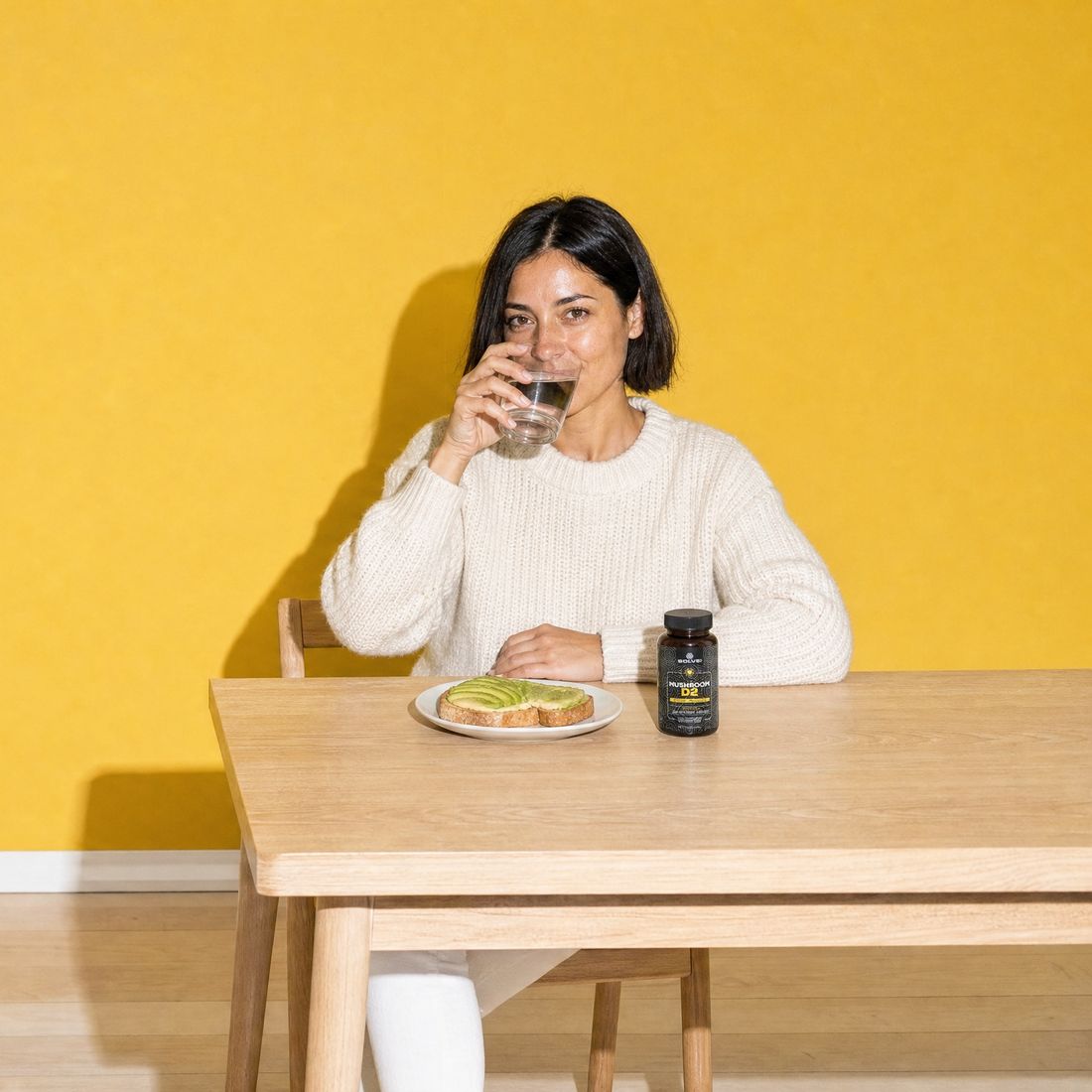 A woman drinking a glass of water at an oak table against a yellow wall, the jar of Vitamin D2 in front of her