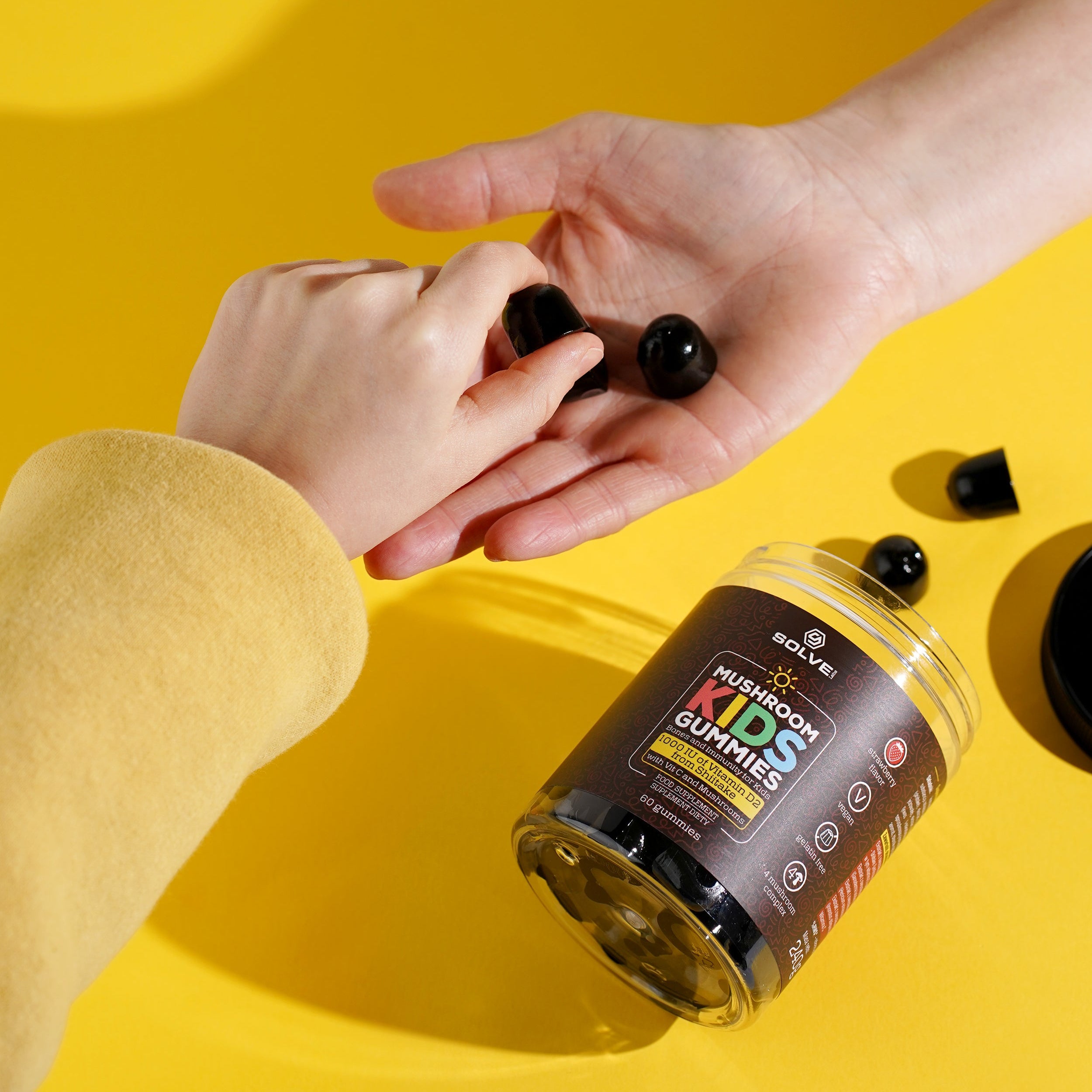 Person holding black gummy bears with a container labeled 'KIDS GUMMIES' on a yellow background