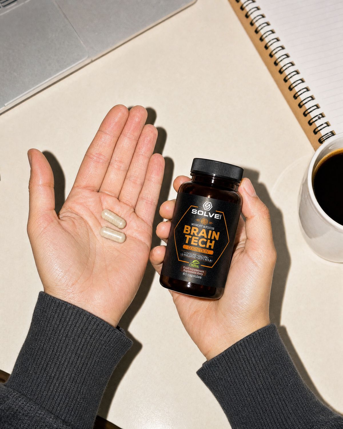 Two Brain Tech capsules in an open palm beside the jar, a laptop and a mug of coffee