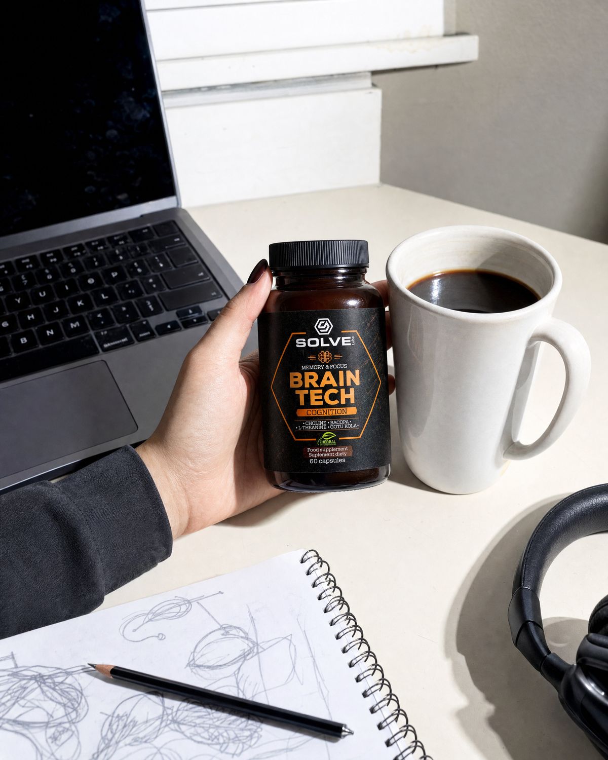 A hand setting the jar of Brain Tech down beside a mug of coffee, a laptop and a sketchbook