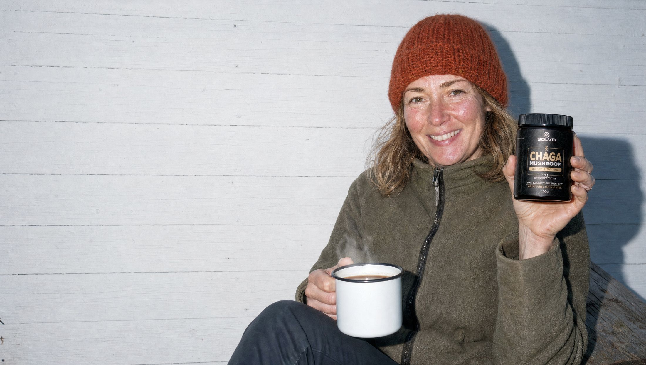 A woman in a wool hat sitting on a cabin step with a steaming mug, holding up a jar of Solve Labs Chaga powder