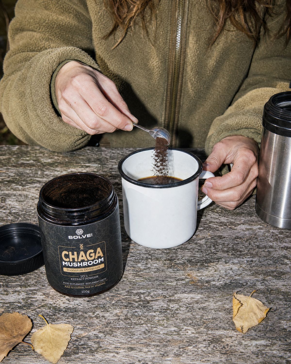 A spoon of chaga powder falling into an enamel mug of coffee beside the open jar