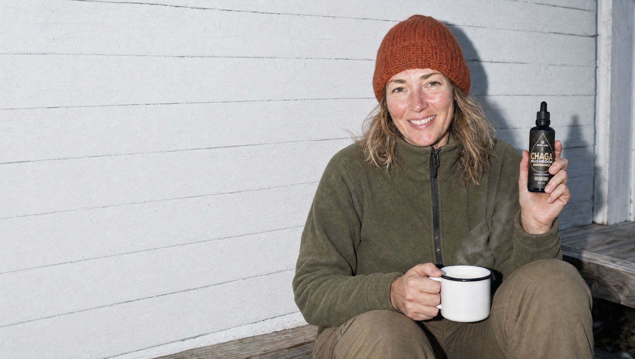 A woman in a wool hat sitting on a cabin step with a steaming mug, holding up a bottle of Solve Labs Chaga drops