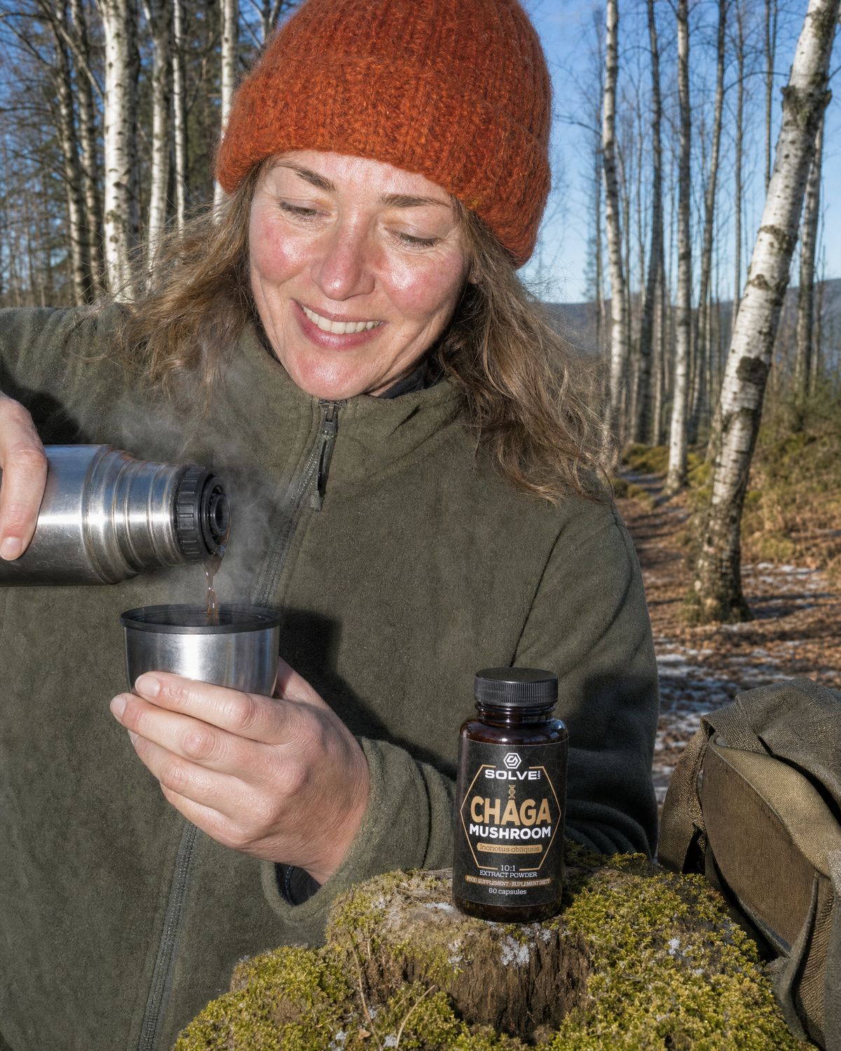 A woman pouring coffee from a flask among birch trees, the jar of Solve Labs Chaga capsules on a tree stump
