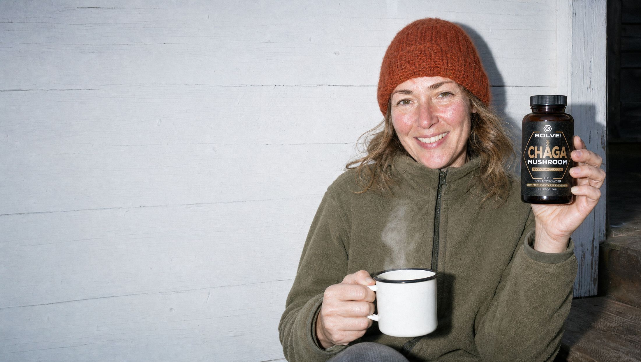 A woman in a wool hat sitting on a cabin step with a steaming mug, holding up a jar of Solve Labs Chaga capsules