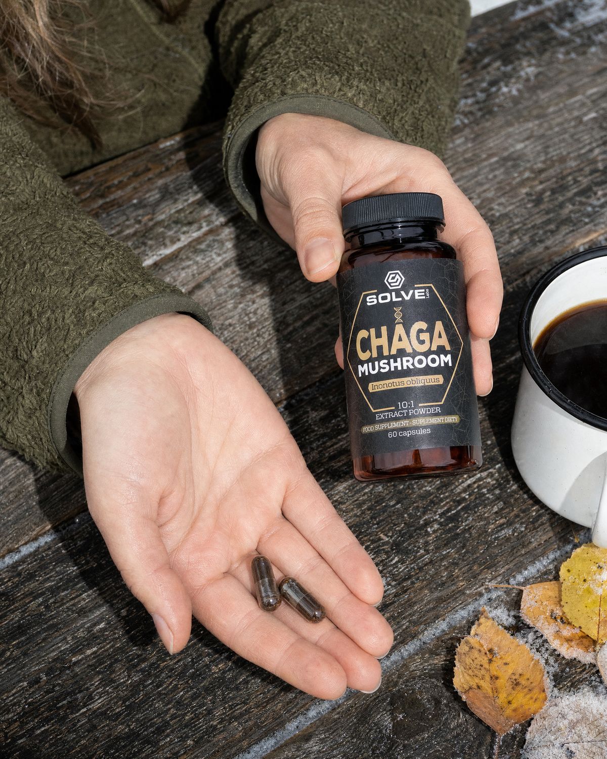 Two chaga capsules in an open palm beside the jar and an enamel mug of coffee