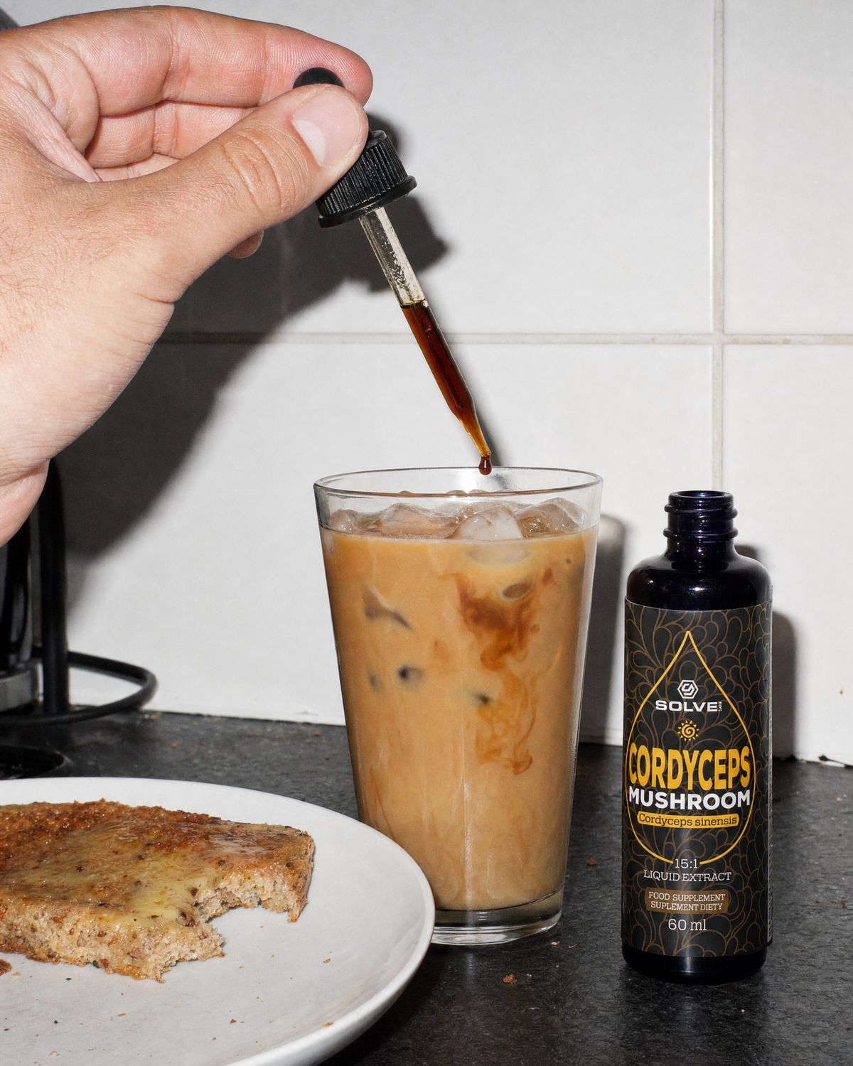 A dropper of cordyceps liquid extract over a glass of iced coffee beside the bottle