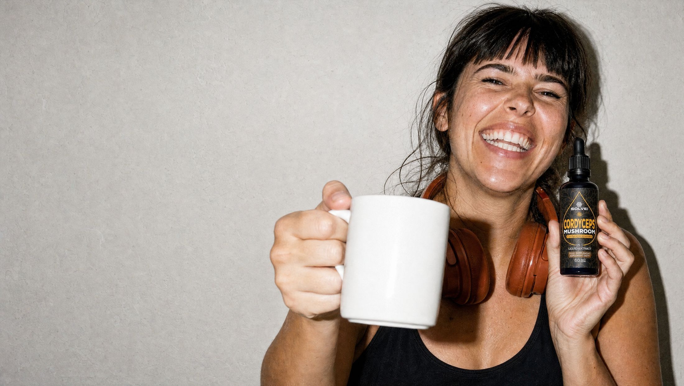 A woman laughing, raising a mug of coffee and holding a bottle of Solve Labs Cordyceps drops