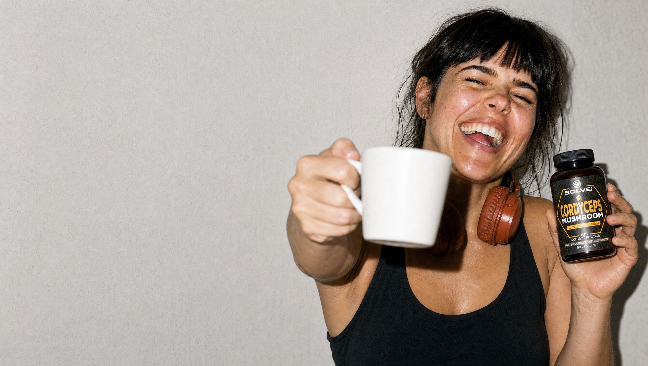A woman laughing, raising a mug of coffee and holding a jar of Solve Labs Cordyceps capsules