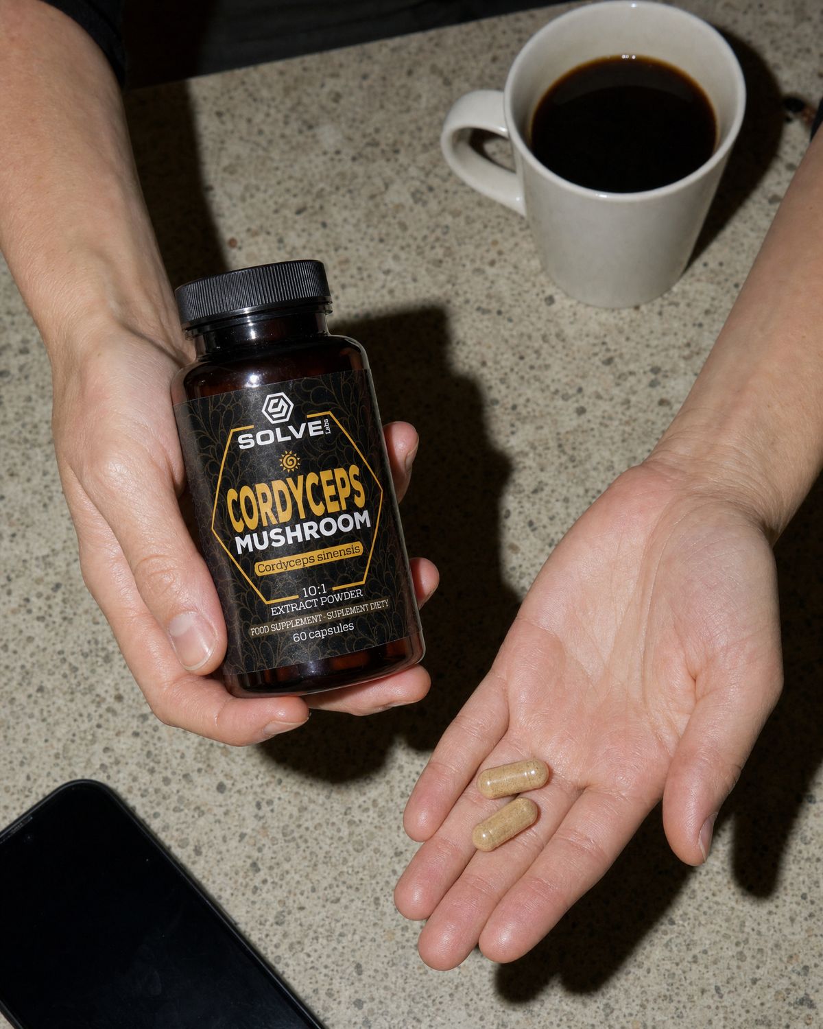 Two cordyceps capsules in an open palm beside the jar and a mug of coffee