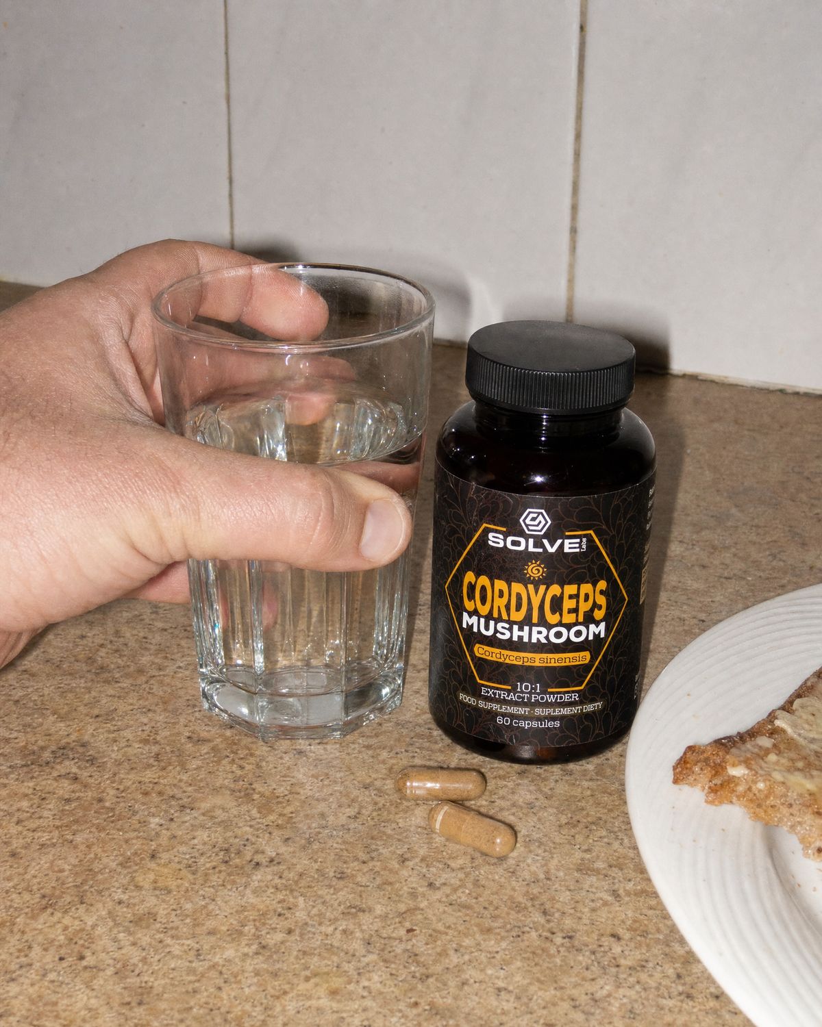 Two cordyceps capsules on a kitchen counter beside a glass of water and the jar