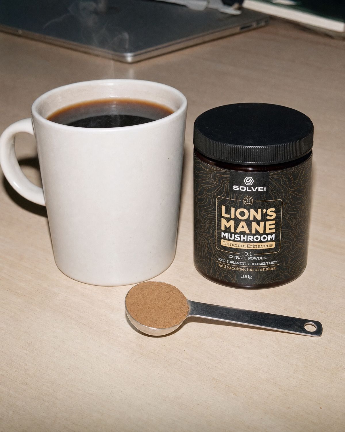A mug of black coffee on a desk, a spoon of lion&rsquo;s mane powder in front of it, the jar beside it