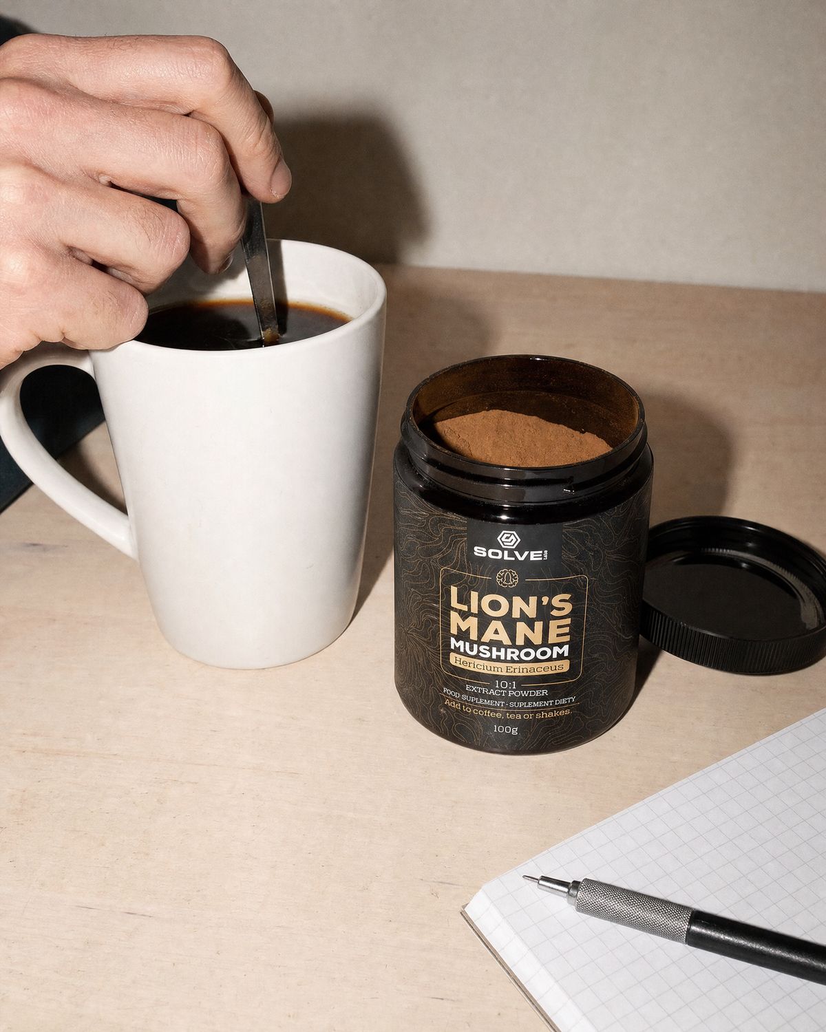A hand stirring a mug of coffee on a desk beside the open jar of Solve Labs Lion&rsquo;s Mane