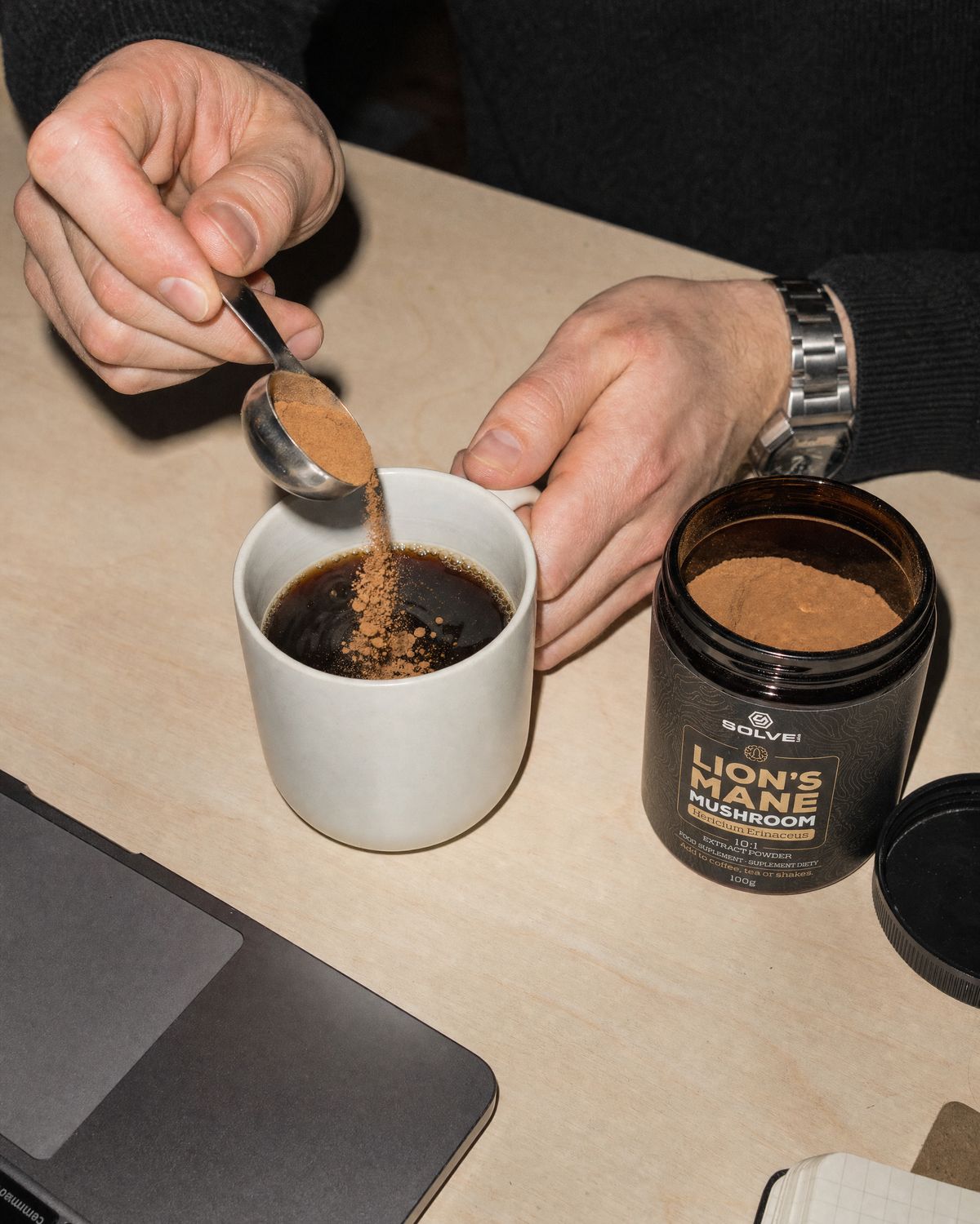 A spoon of lion&rsquo;s mane powder falling into a mug of coffee on a desk beside the open jar
