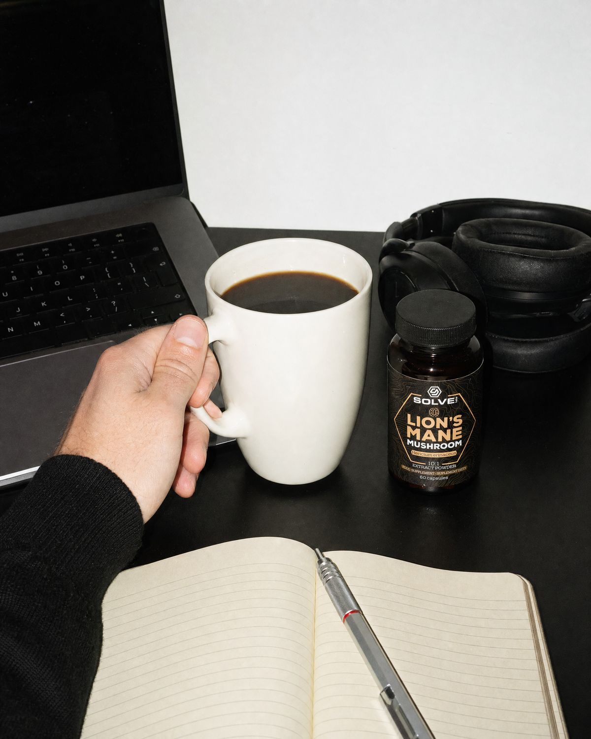 A working desk with a laptop, a notebook, a mug of coffee and the jar of Solve Labs Lion&rsquo;s Mane capsules