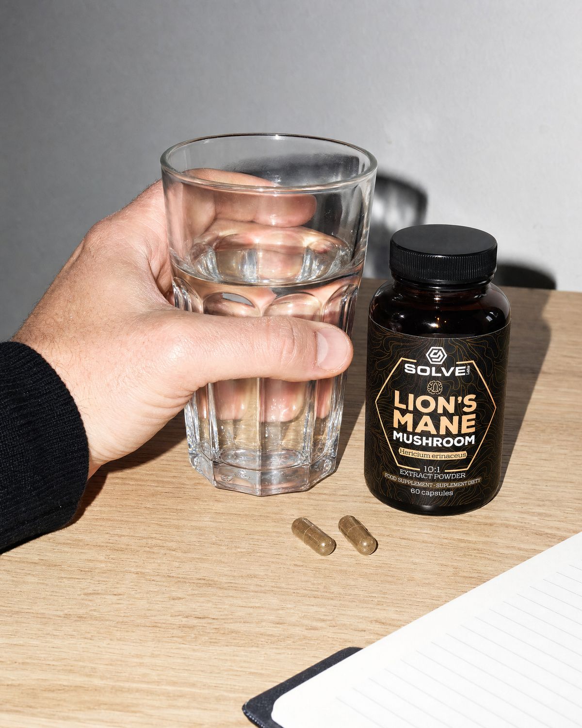 Two lion&rsquo;s mane capsules on a desk beside a glass of water and the jar