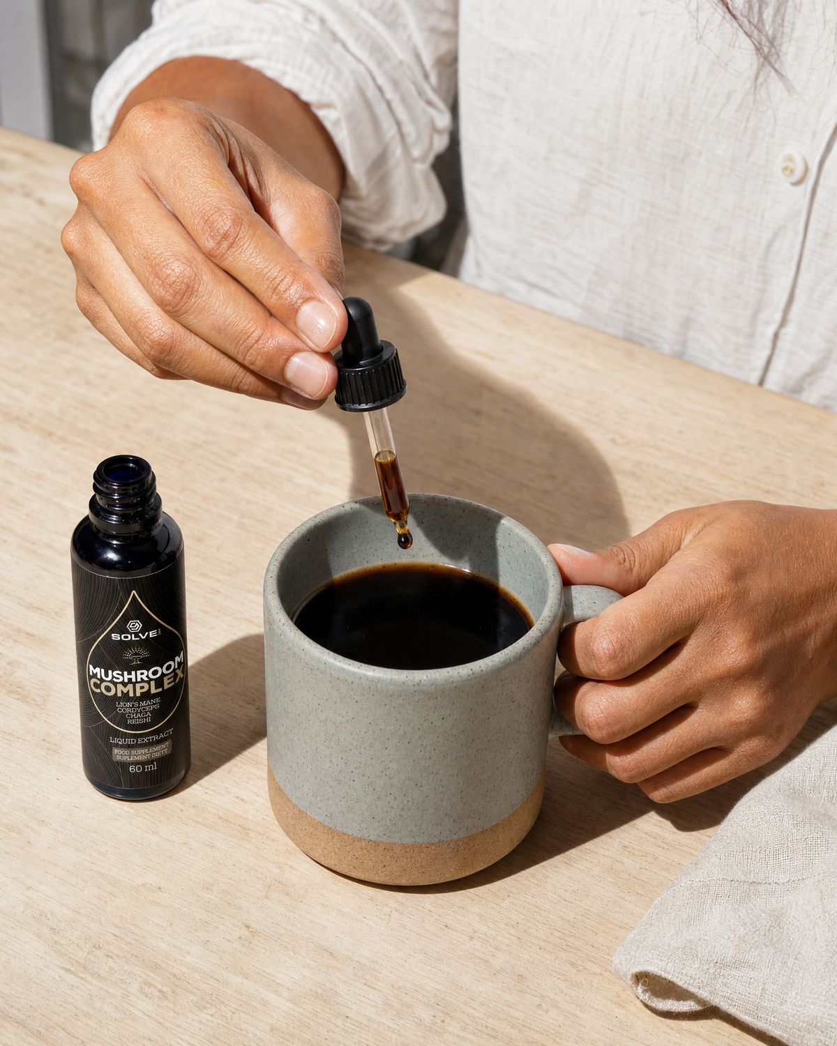 A dropper of Mushroom Complex liquid extract over a stoneware mug of coffee beside the open bottle