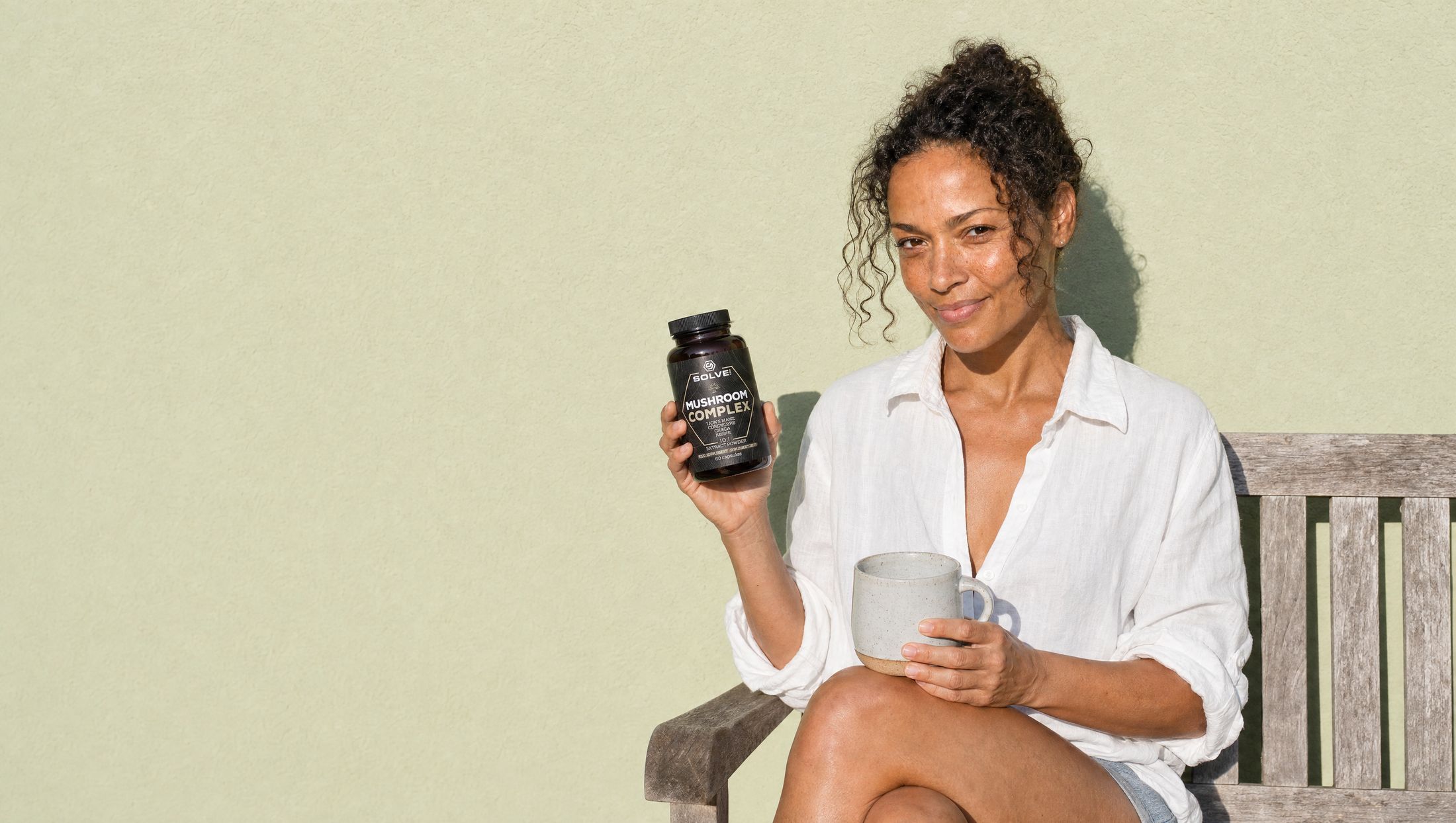 A woman in a white linen shirt on a garden bench with a stoneware mug, holding up a jar of Solve Labs Mushroom Complex capsules