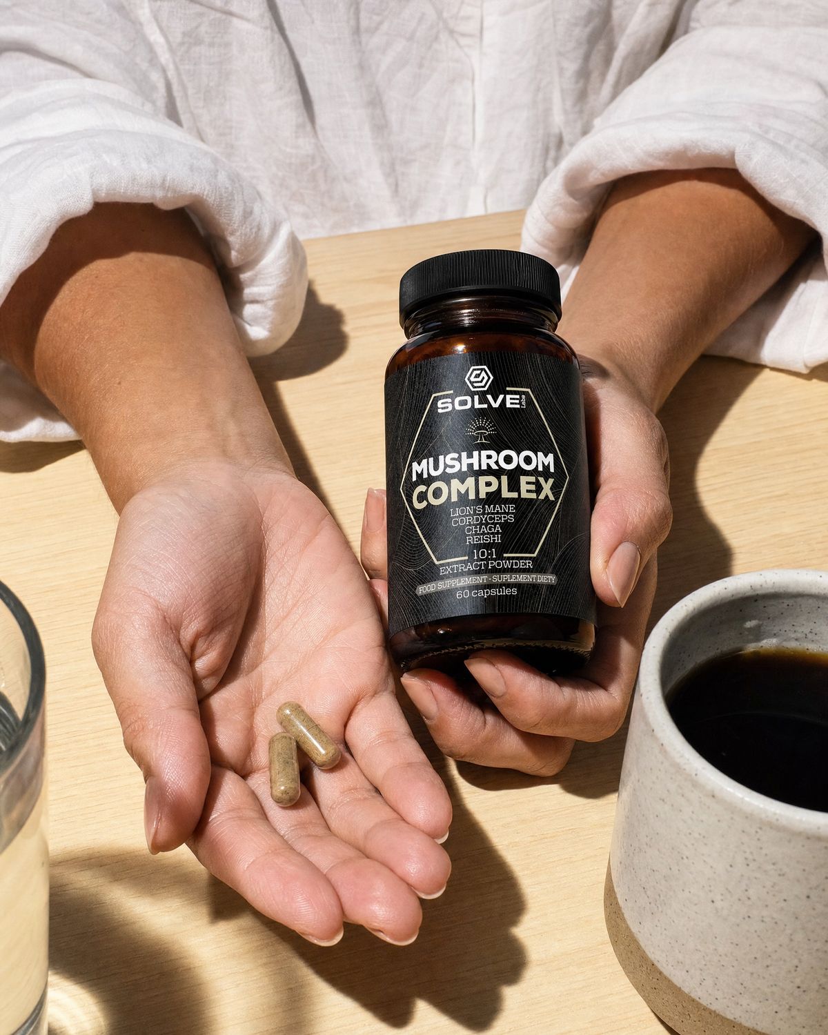 Two Mushroom Complex capsules in an open palm beside the jar, a stoneware mug and a glass of water