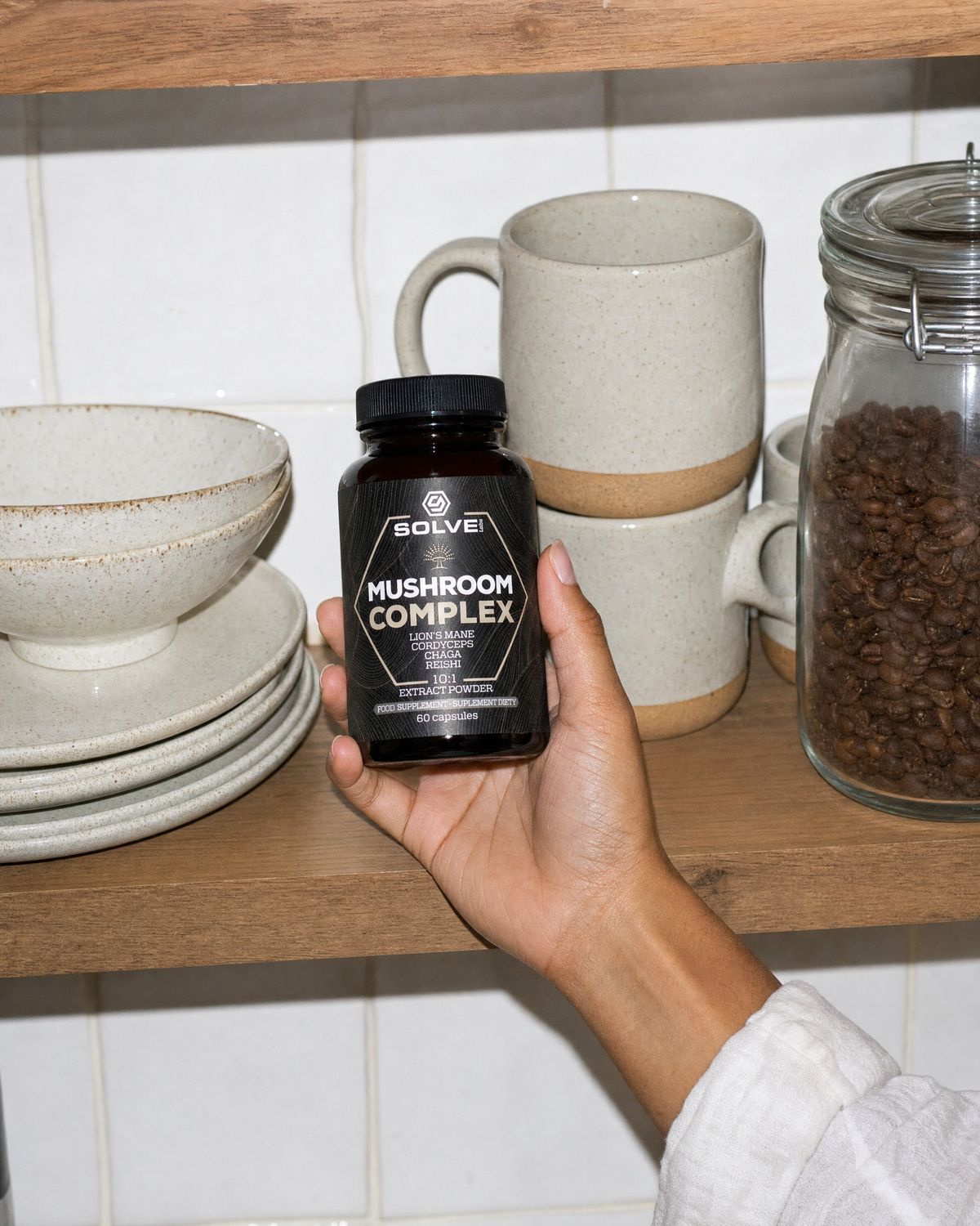 A hand taking the jar of Solve Labs Mushroom Complex capsules from a kitchen shelf of mugs and bowls
