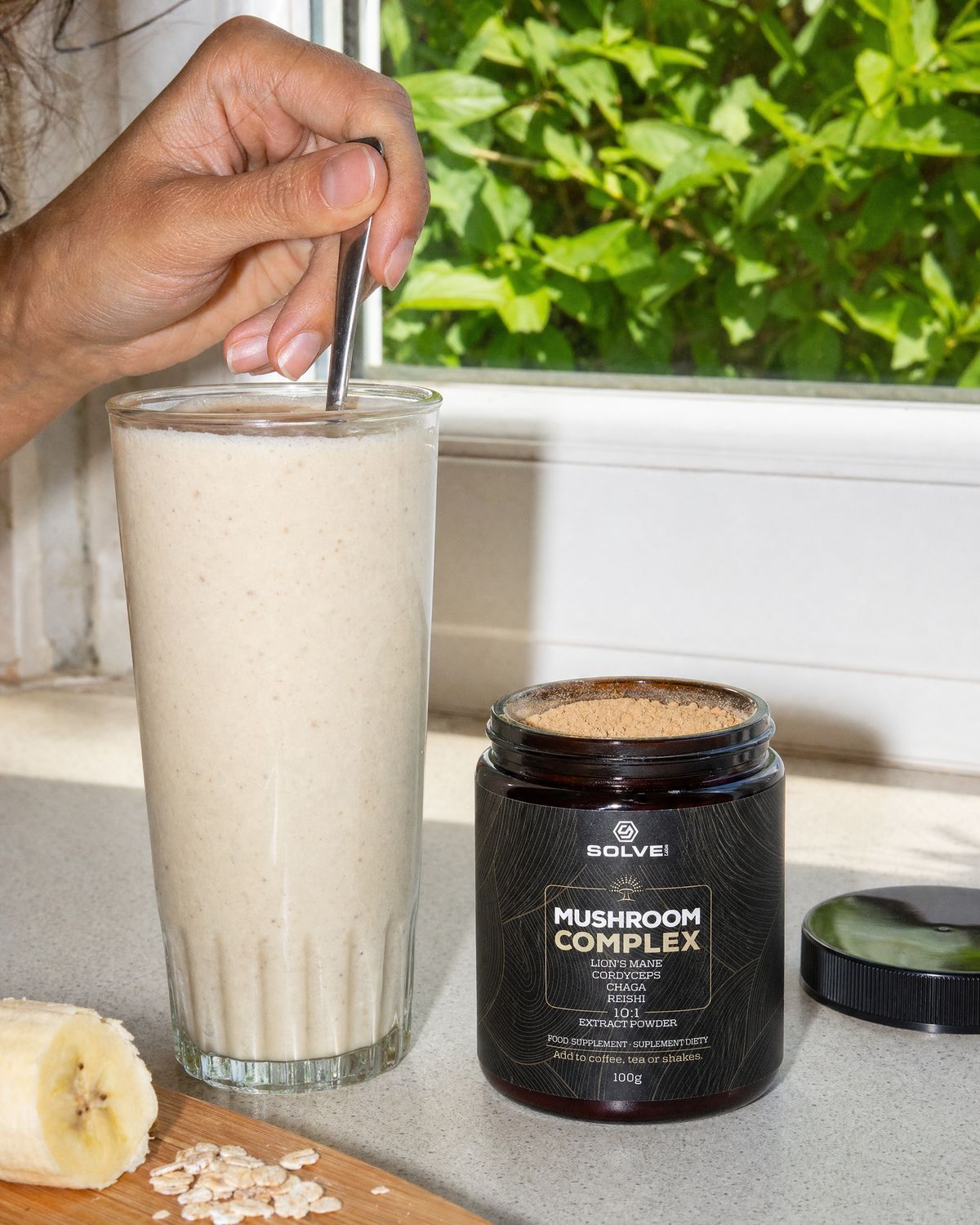 A hand stirring a glass of banana smoothie beside the open jar of Solve Labs Mushroom Complex powder