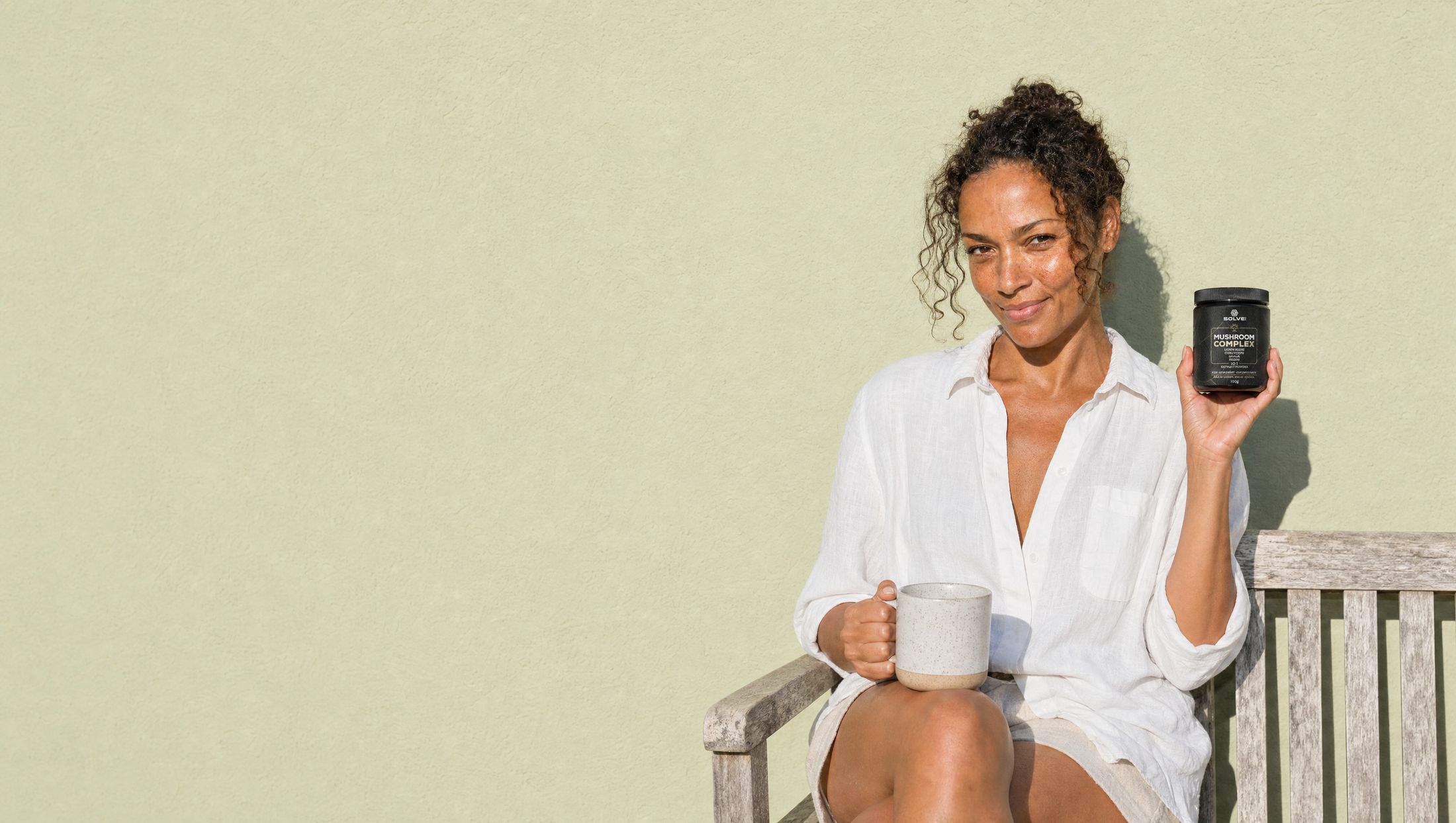 A woman in a white linen shirt on a garden bench with a stoneware mug, holding up a jar of Solve Labs Mushroom Complex powder