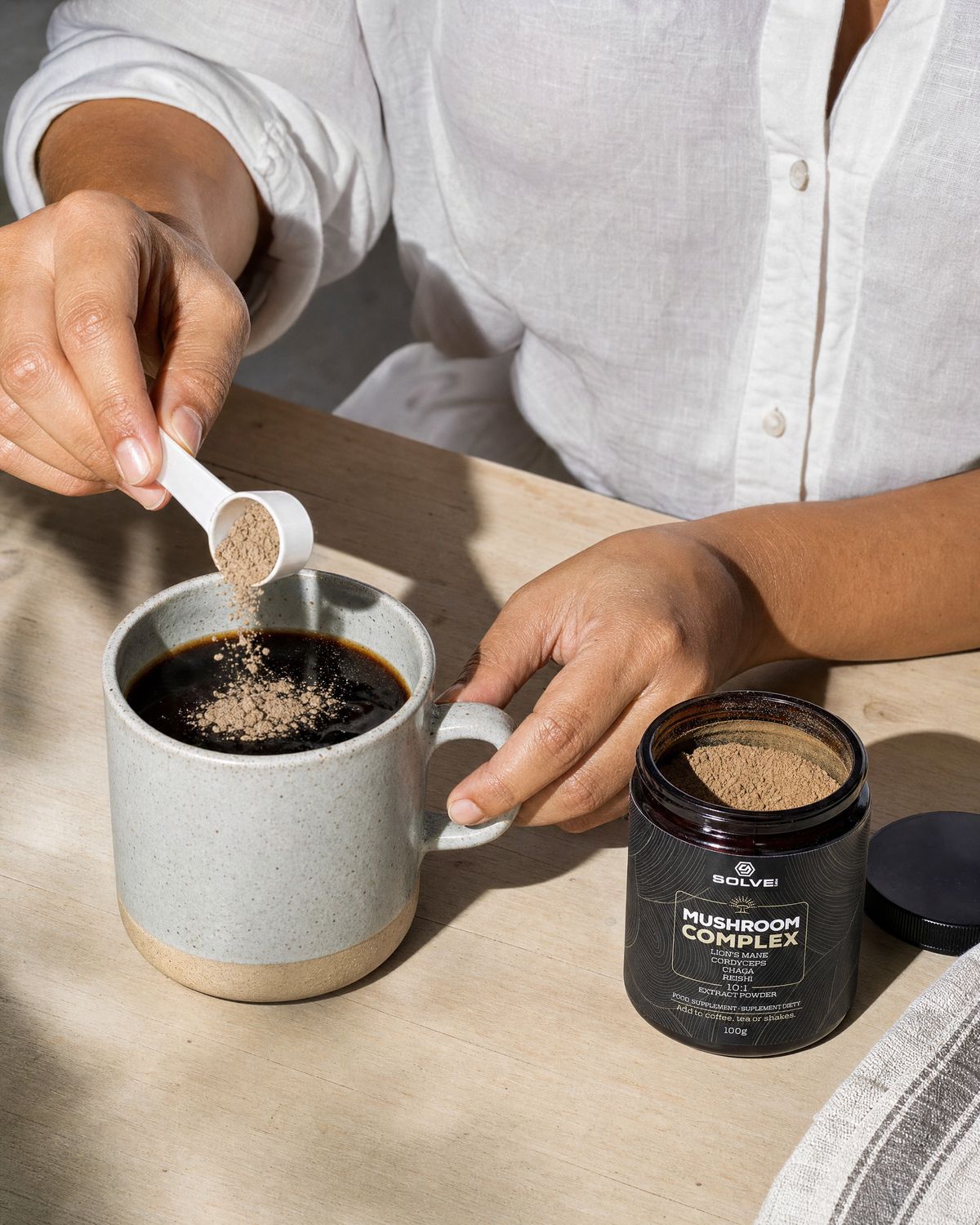 A scoop of Mushroom Complex powder falling into a stoneware mug of coffee beside the open jar