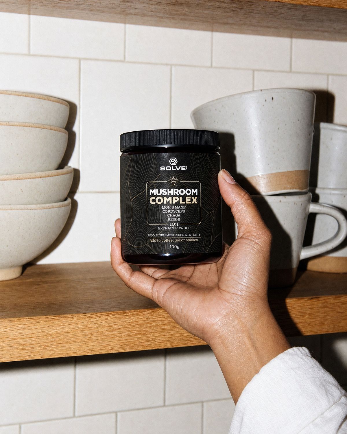 A hand taking the jar of Solve Labs Mushroom Complex powder from a kitchen shelf of mugs and bowls