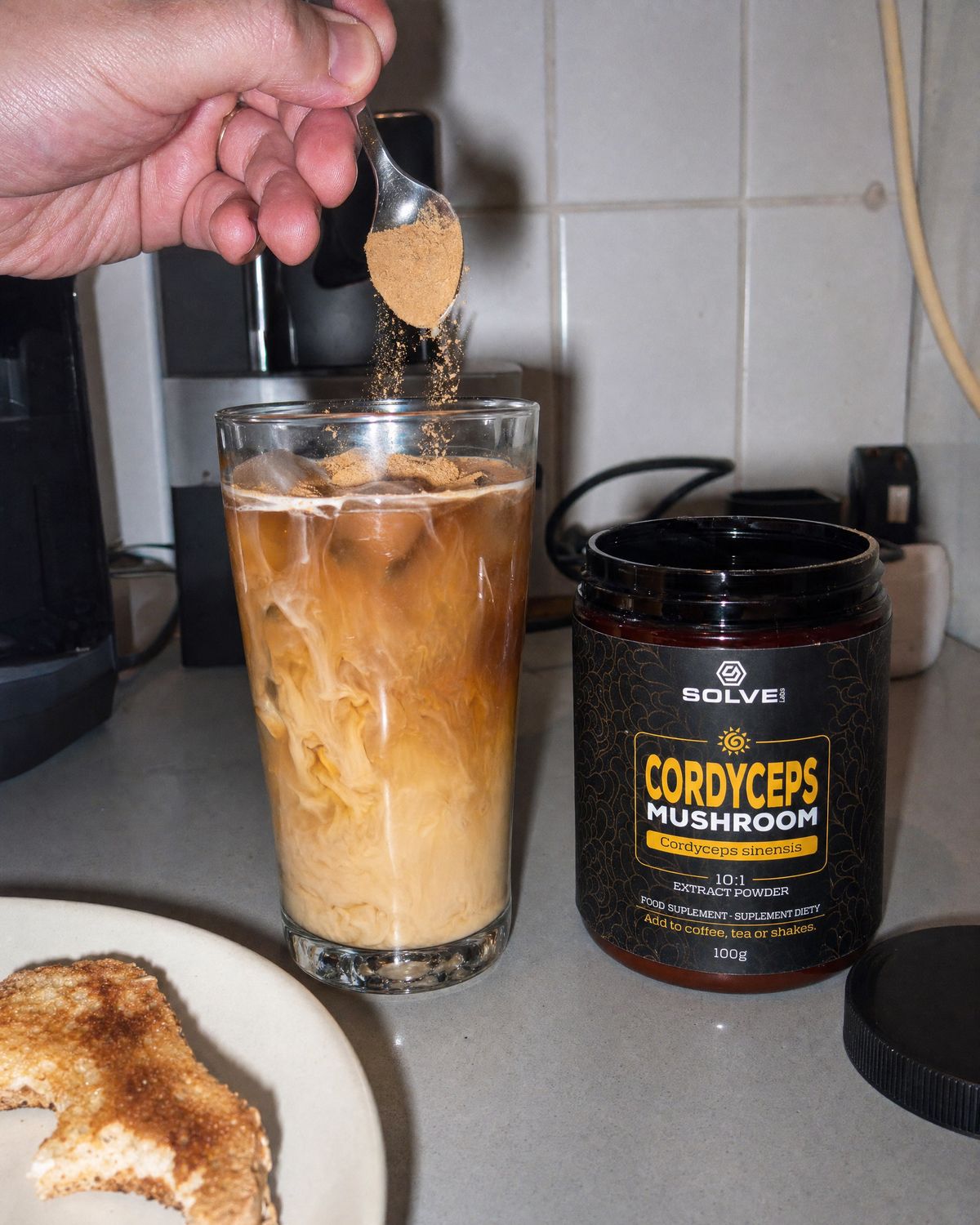 A spoon of cordyceps powder going into a glass of iced coffee beside the jar
