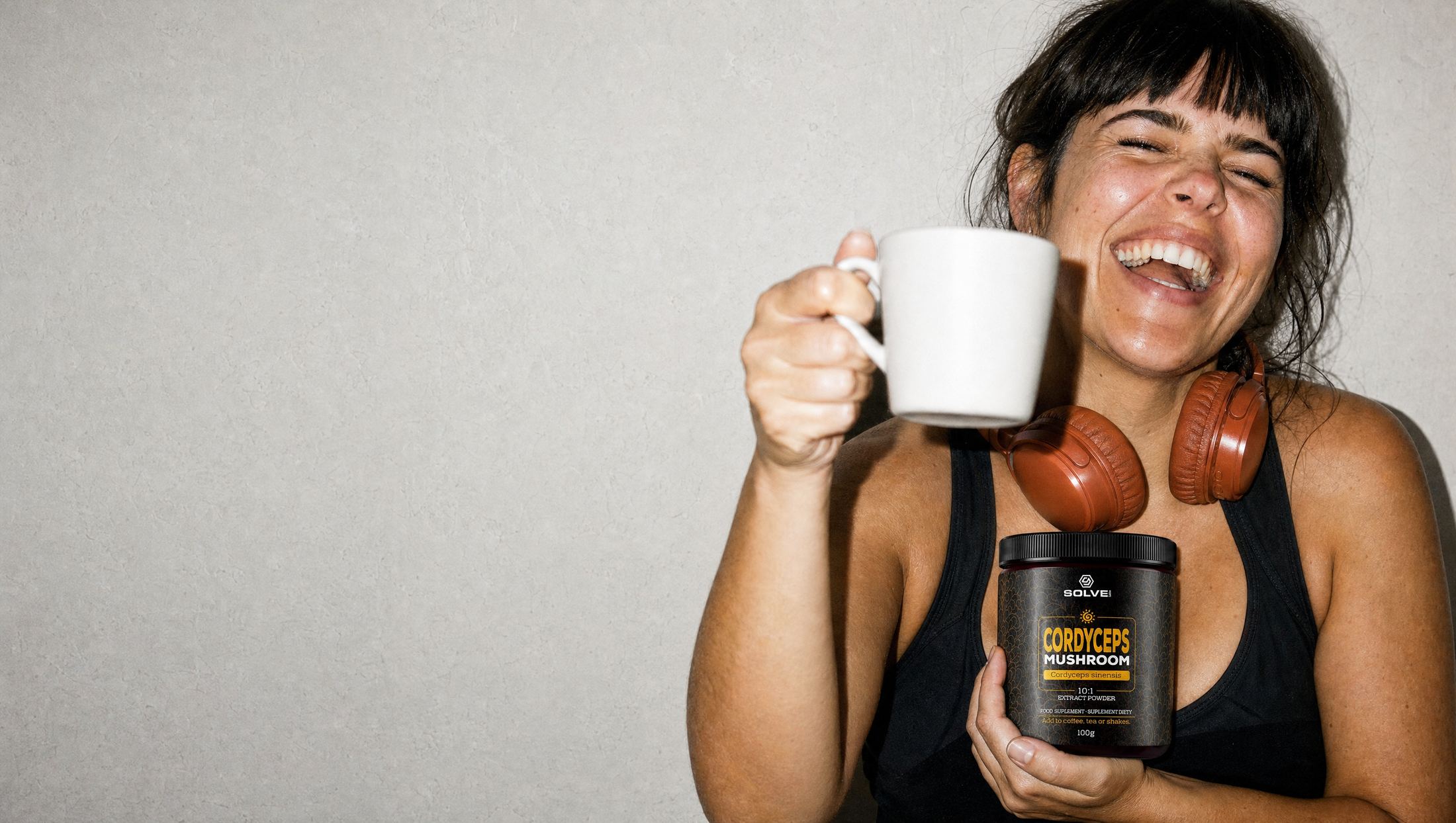 A woman laughing, raising a mug of coffee and holding a jar of Solve Labs Cordyceps powder