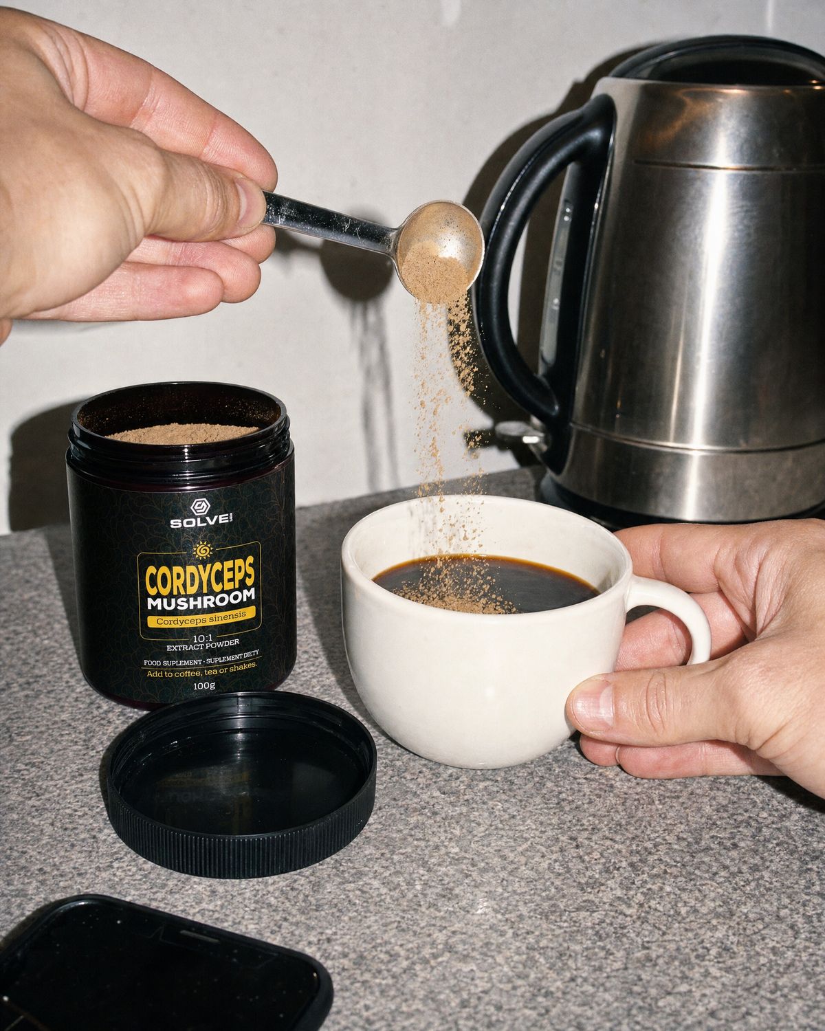 A spoon of cordyceps powder falling into a mug of coffee beside the open jar