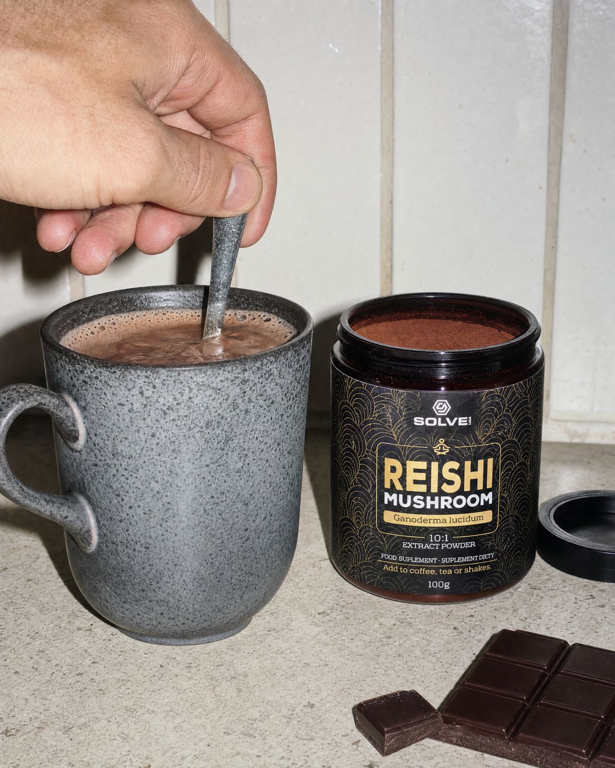 A hand stirring a mug of hot cacao beside the open jar of Solve Labs Reishi
