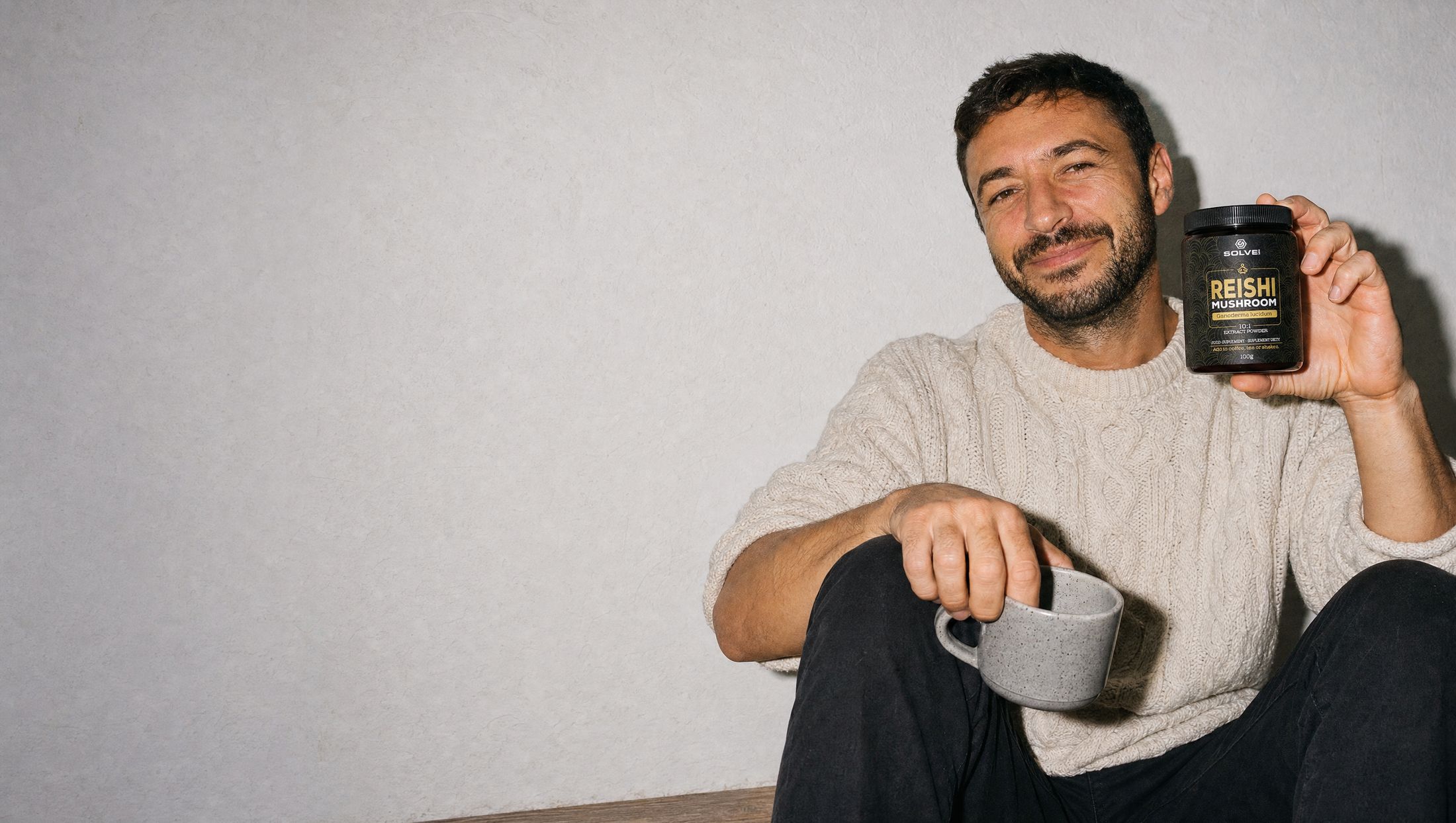 A man sitting against a wall with a mug on his knee and a calm smile, holding up a jar of Solve Labs Reishi powder