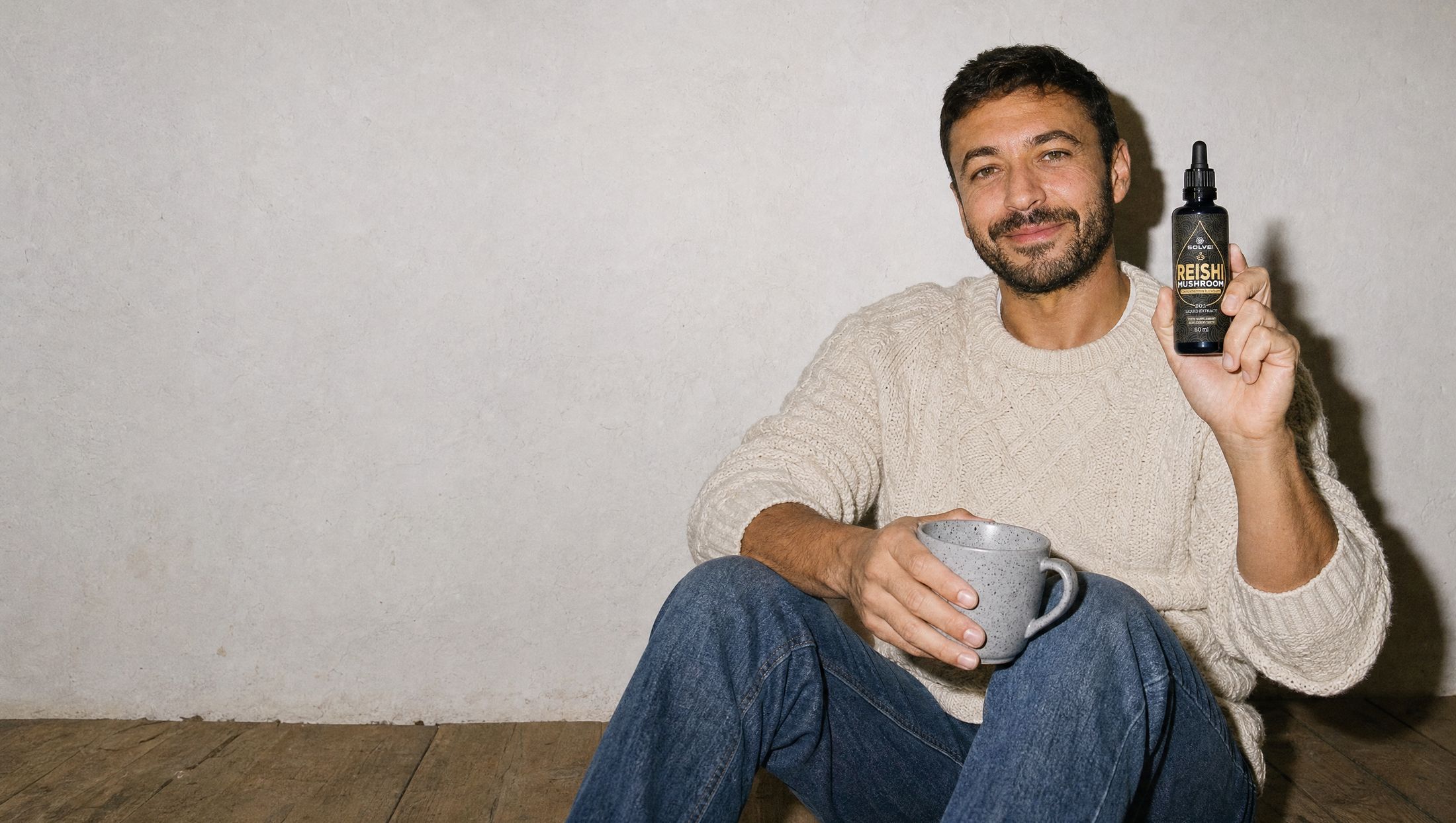 A man sitting against a wall with a mug on his knee and a calm smile, holding up a bottle of Solve Labs Reishi drops
