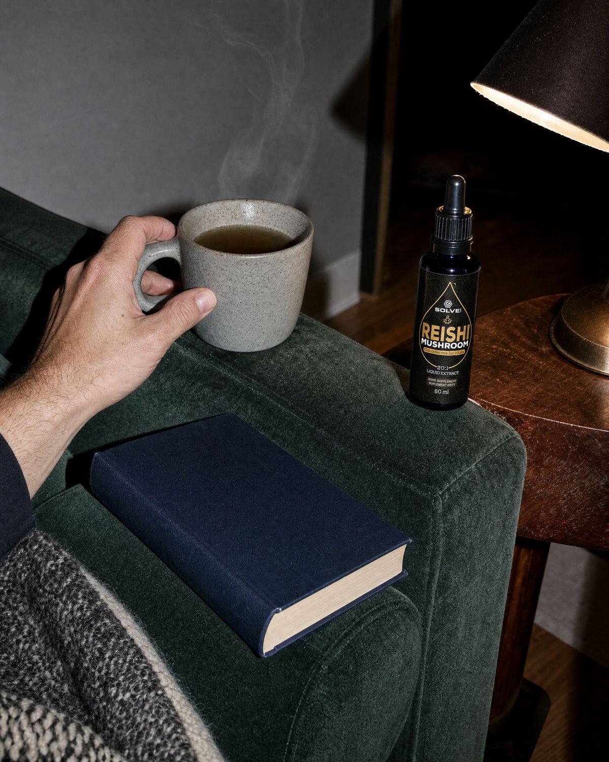 A mug of herbal tea on the arm of a sofa beside a book, the bottle of Solve Labs Reishi drops on a side table