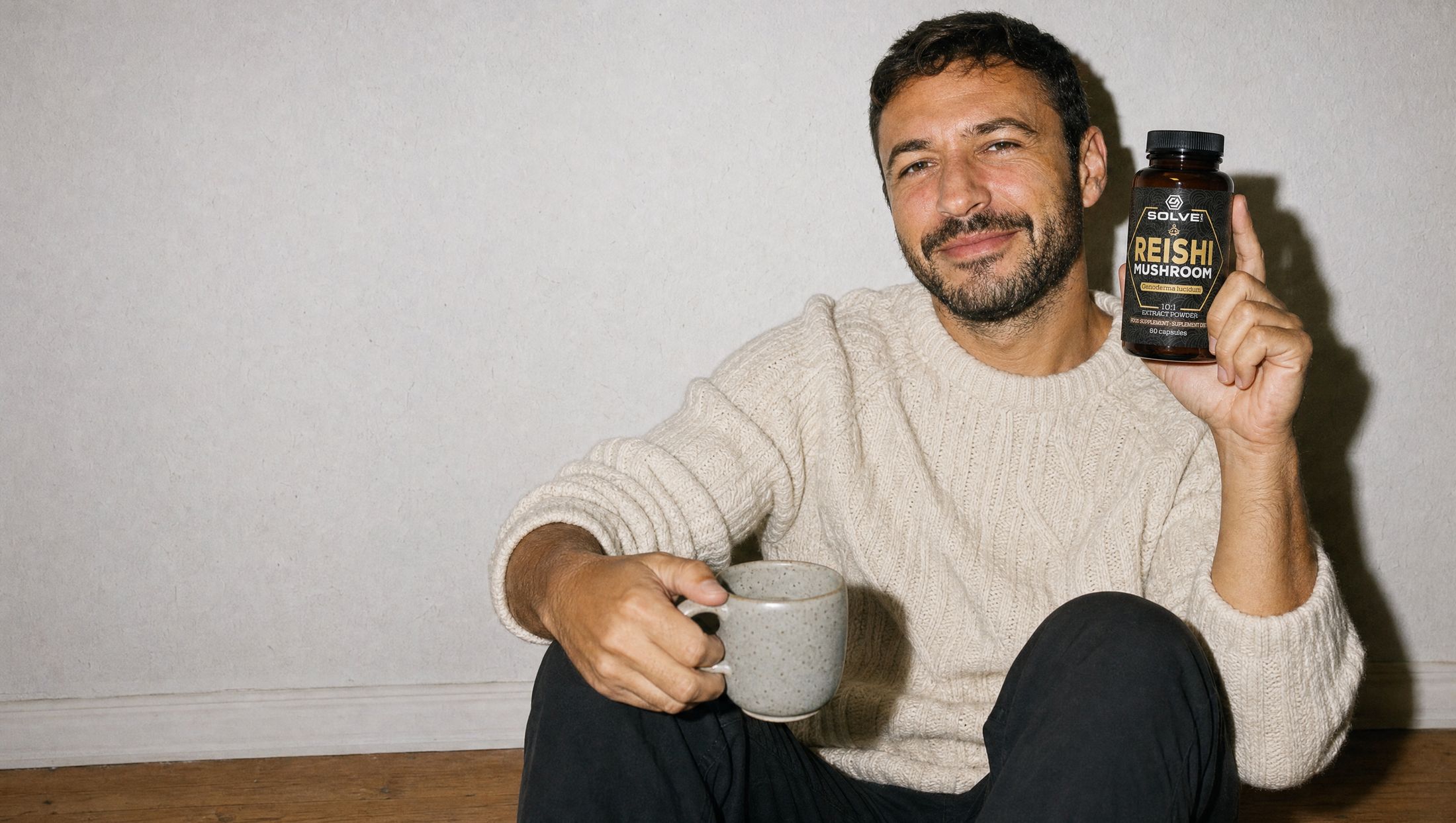 A man sitting against a wall with a mug on his knee and a calm smile, holding up a jar of Solve Labs Reishi capsules