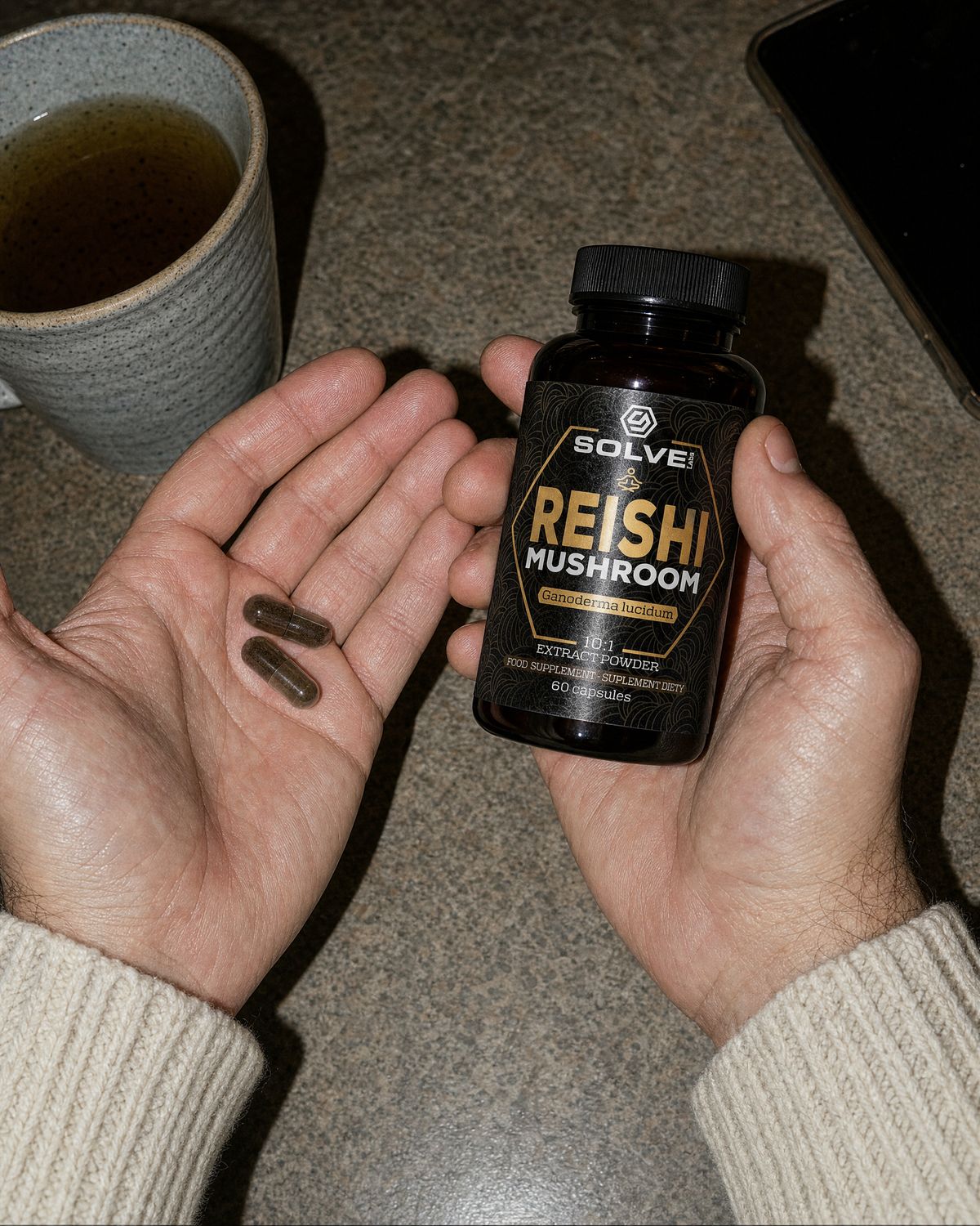 Two reishi capsules in an open palm beside the jar and a mug of herbal tea