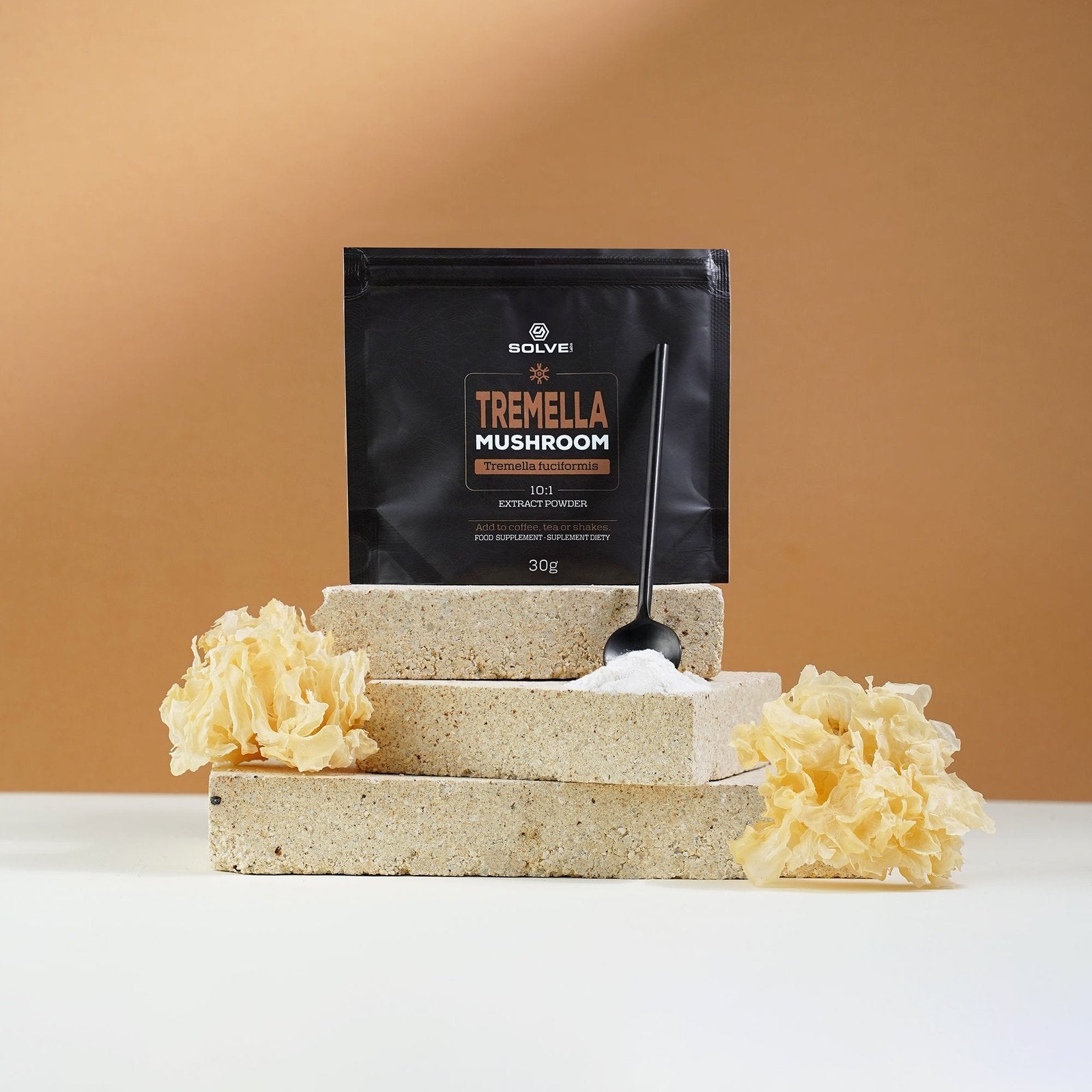 Der Beutel Solve Labs Tremella-Pulver steht auf hellen Steinplatten, davor ein Löffel des weißen Pulvers, auf beiden Seiten getrockneter Schneepilz
