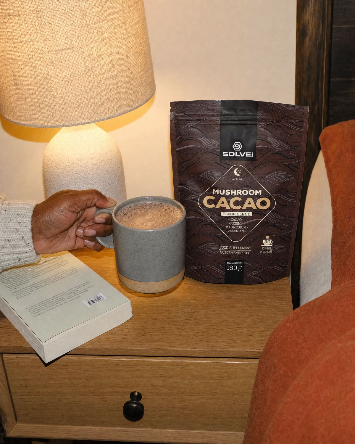 A hand setting a mug of cacao down on a bedside table beside the pouch, a lamp and a book