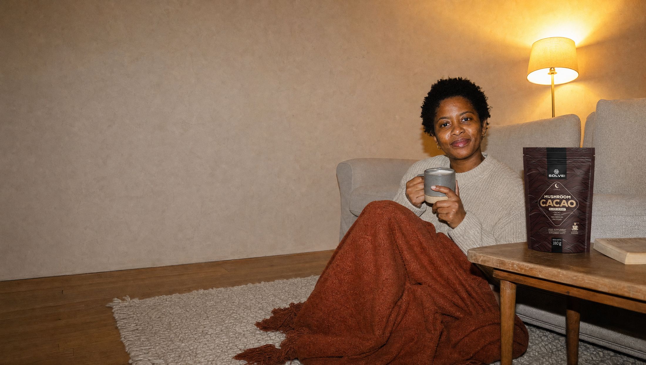 A woman curled up on a rug by the sofa in the evening with a mug of cacao, the pouch of Solve Labs Mushroom Cacao on the table beside her