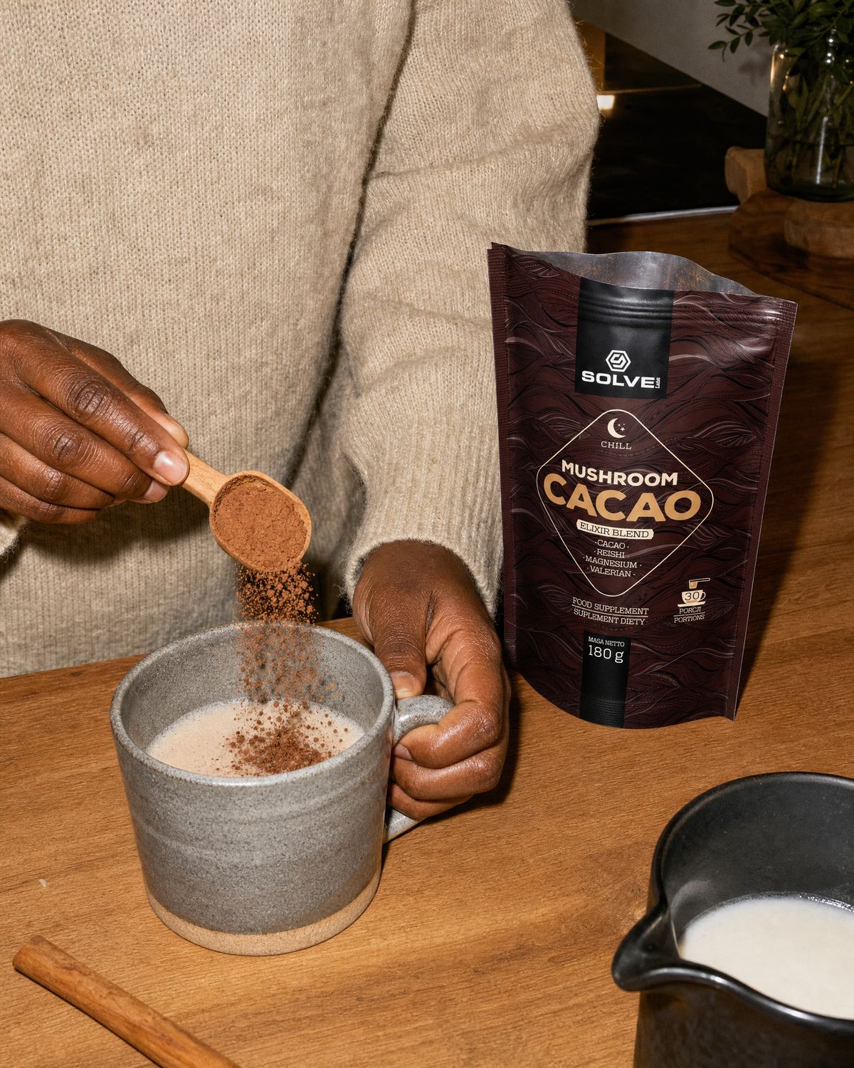 A wooden scoop of cacao powder falling into a mug of hot milk beside the open pouch