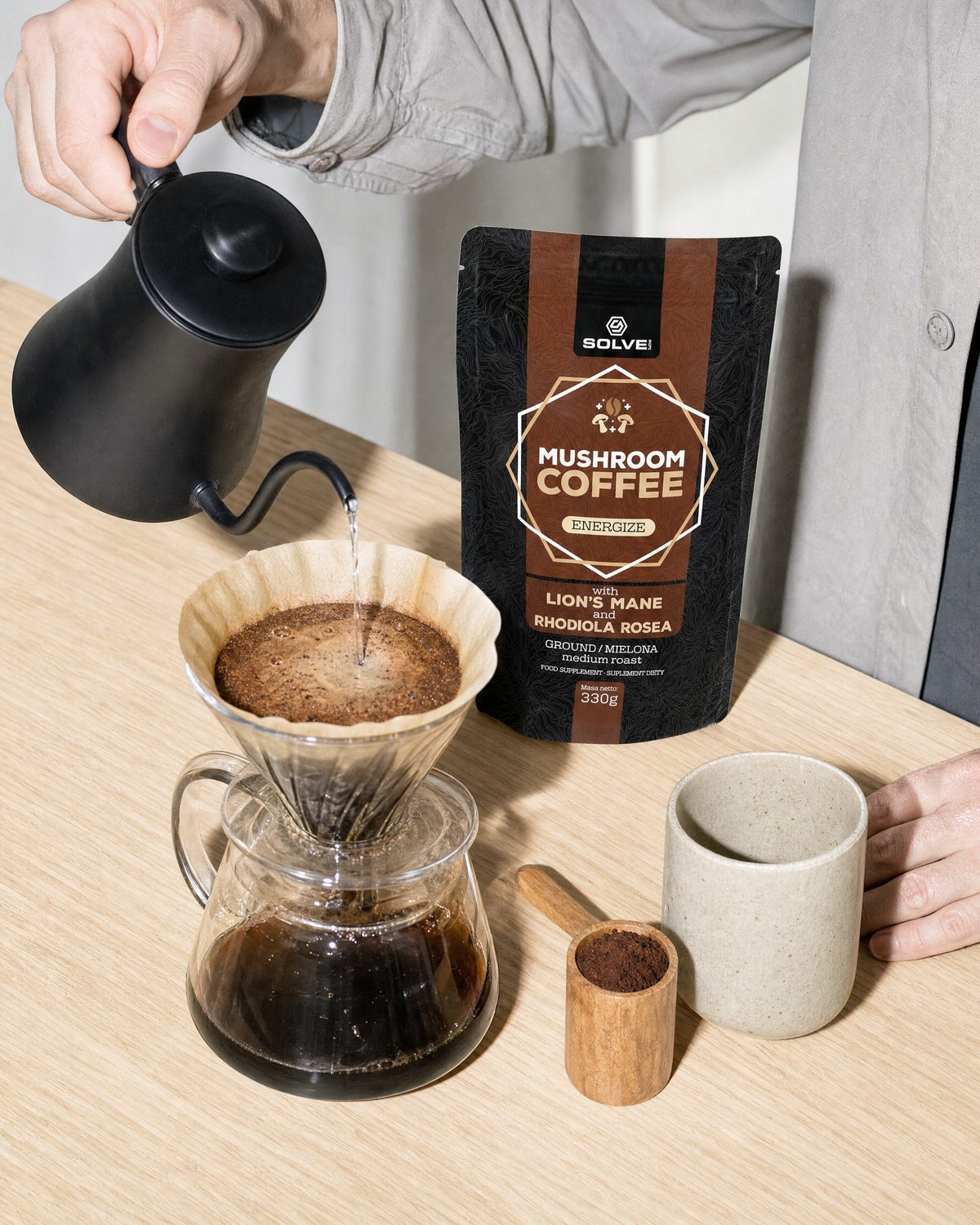 Hands pouring water from a kettle over a pour-over carafe, a bag of Mushroom Coffee with Lion&rsquo;s Mane and a stoneware cup beside it